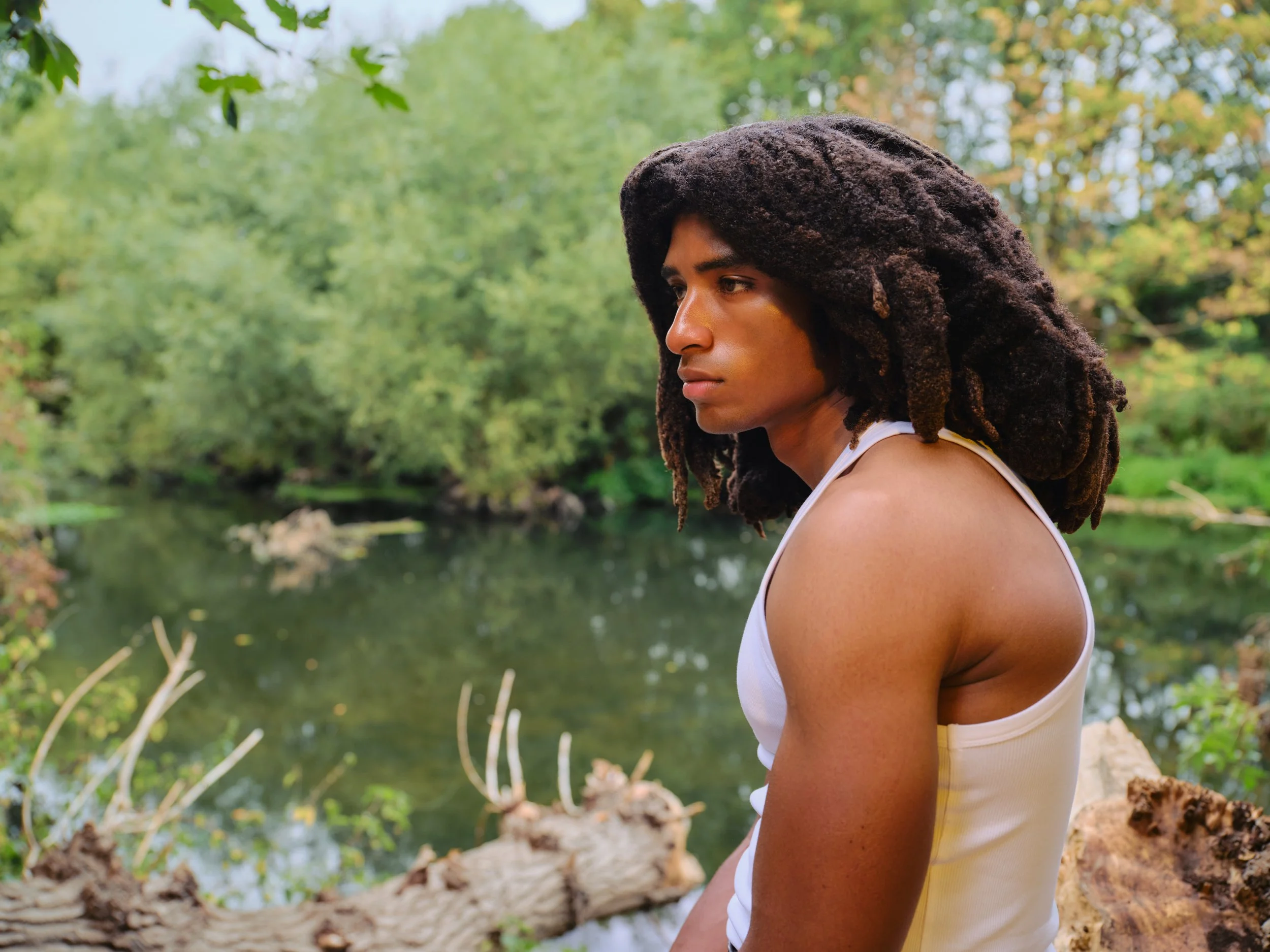 Profile of a woman with dark curly locs sitting outdoors by a body of water, surrounded by green trees, wearing a white tank top.