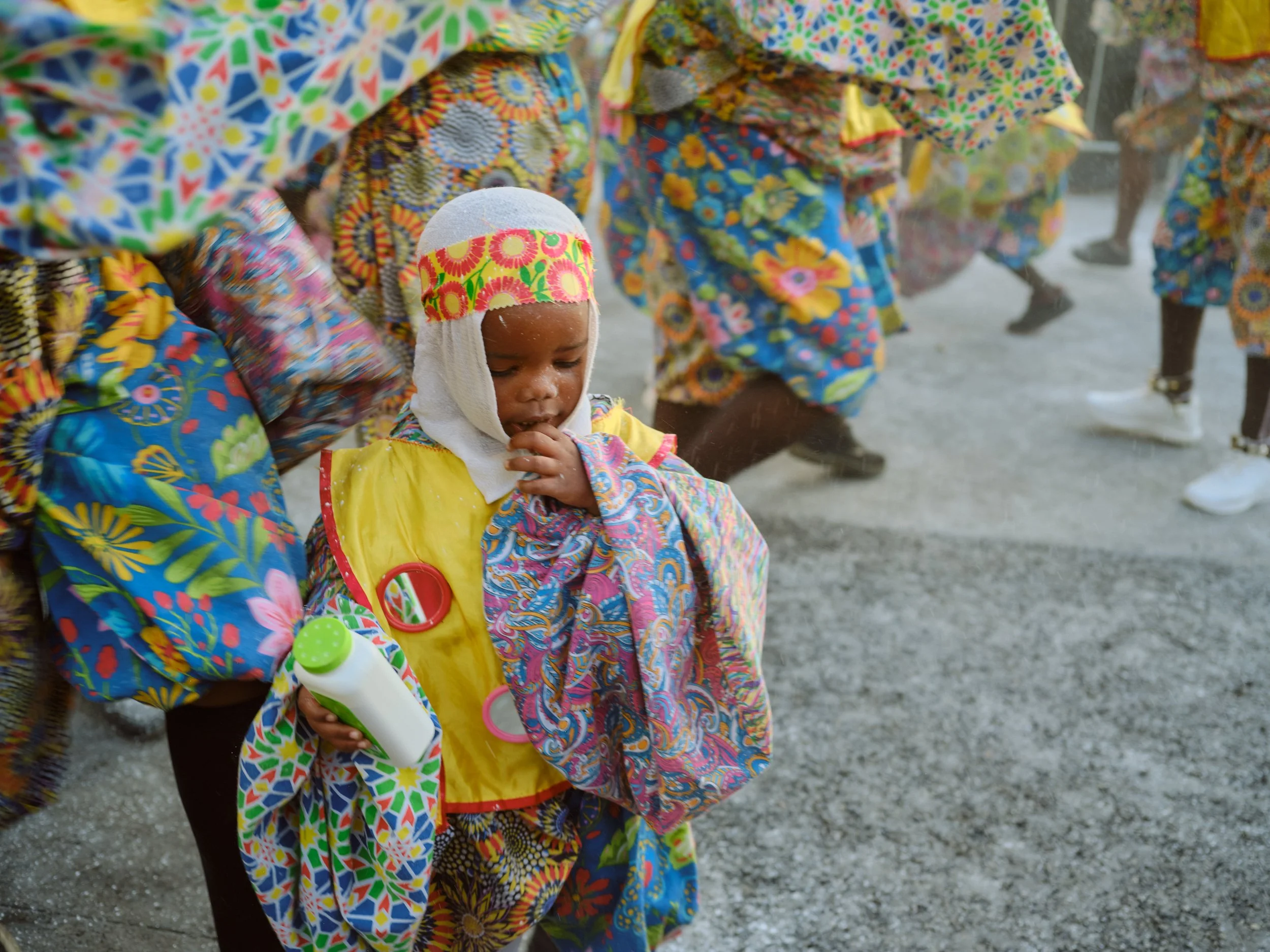 A young child wearing a yellow raincoat with colorful, patterned clothing underneath, holding a white water bottle with a green cap, surrounded by people in vibrant, colorful clothing with floral patterns.