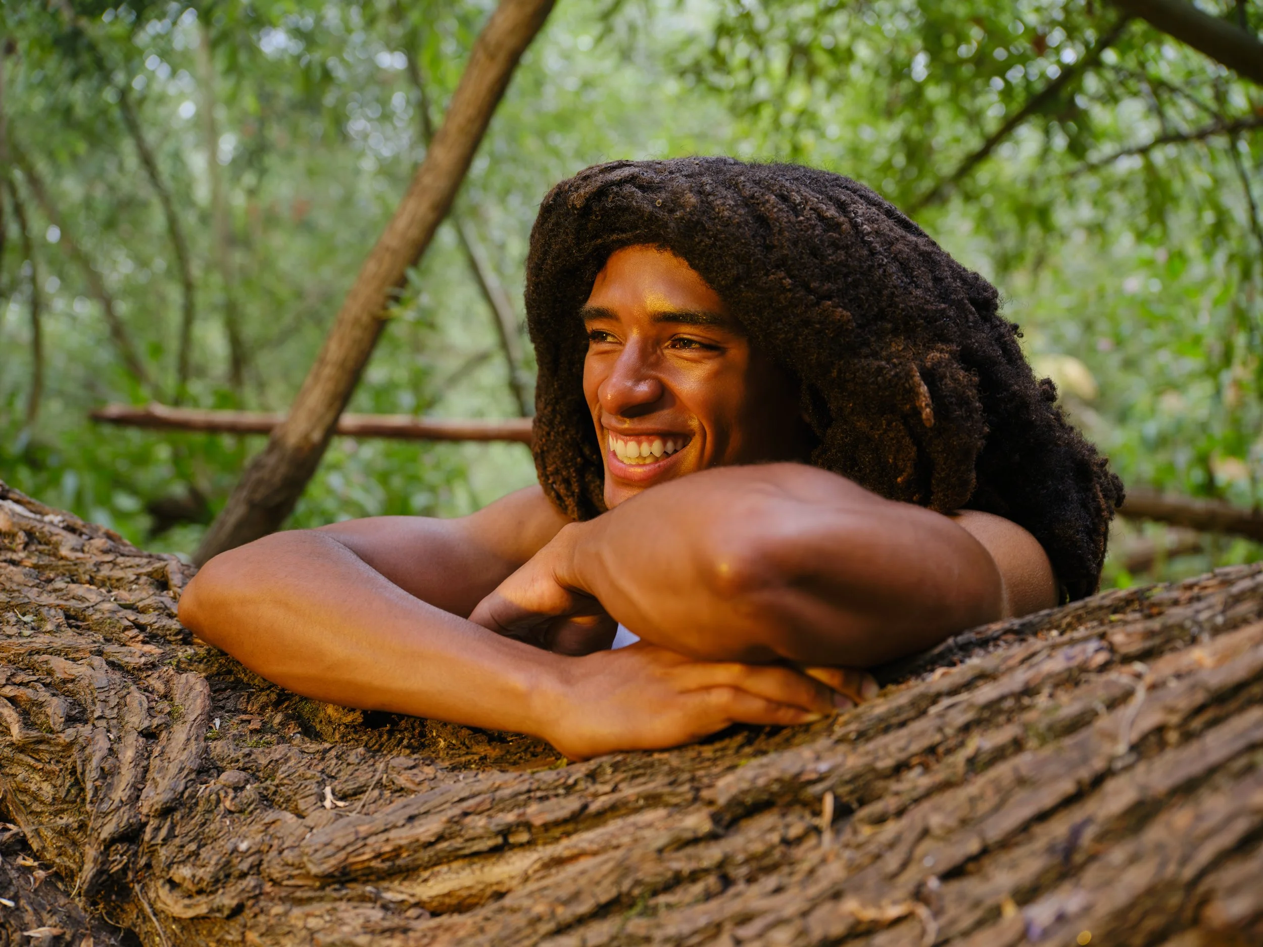 A person with curly dark hair smiling and resting their head on folded arms on a tree branch in a lush green forest.