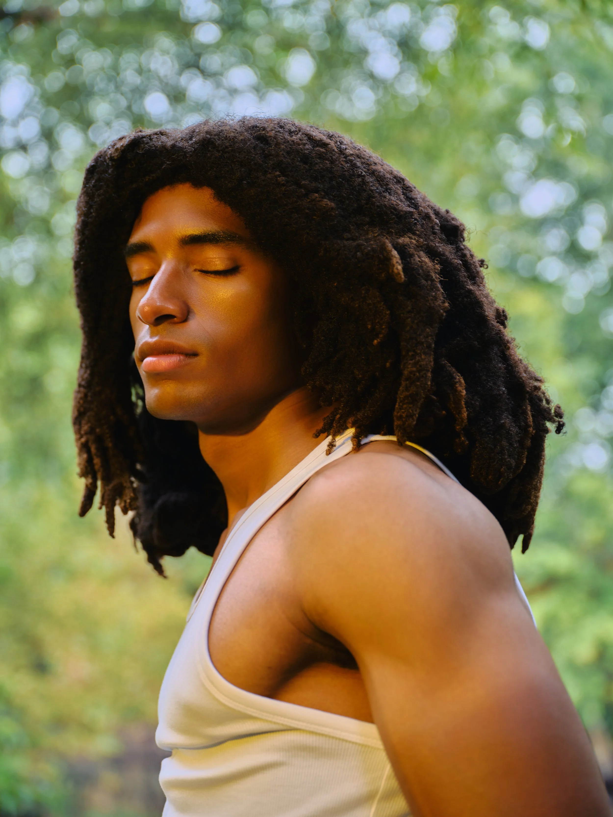 A woman with dark curly dreadlocks relaxing outdoors with eyes closed, wearing a sleeveless white top, with a background of green trees and bokeh sunlight.