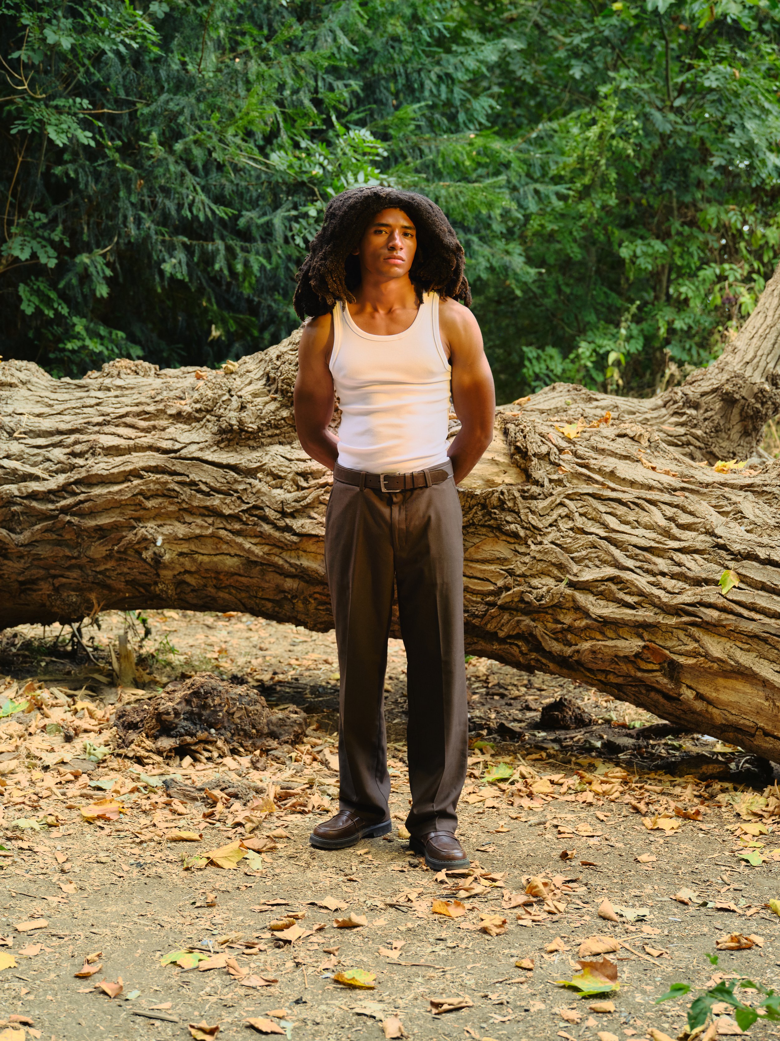 A young man with dark curly hair, wearing a white sleeveless shirt and dark pants, stands with hands behind his back in front of a large fallen tree trunk in a forest with green foliage.