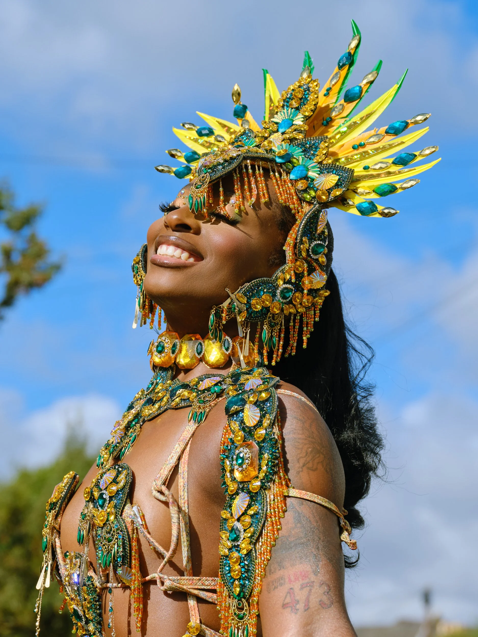 A person wearing an elaborate and colorful headdress and jewelry, smiling with closed eyes, against a blue sky background.