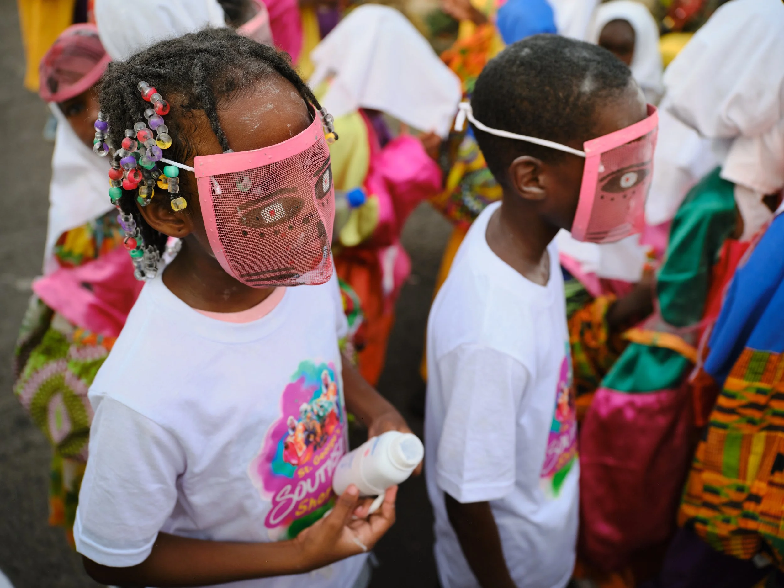 Children wearing pink face masks with eye and lip designs, dressed in colorful clothing, participating in an outdoor event.