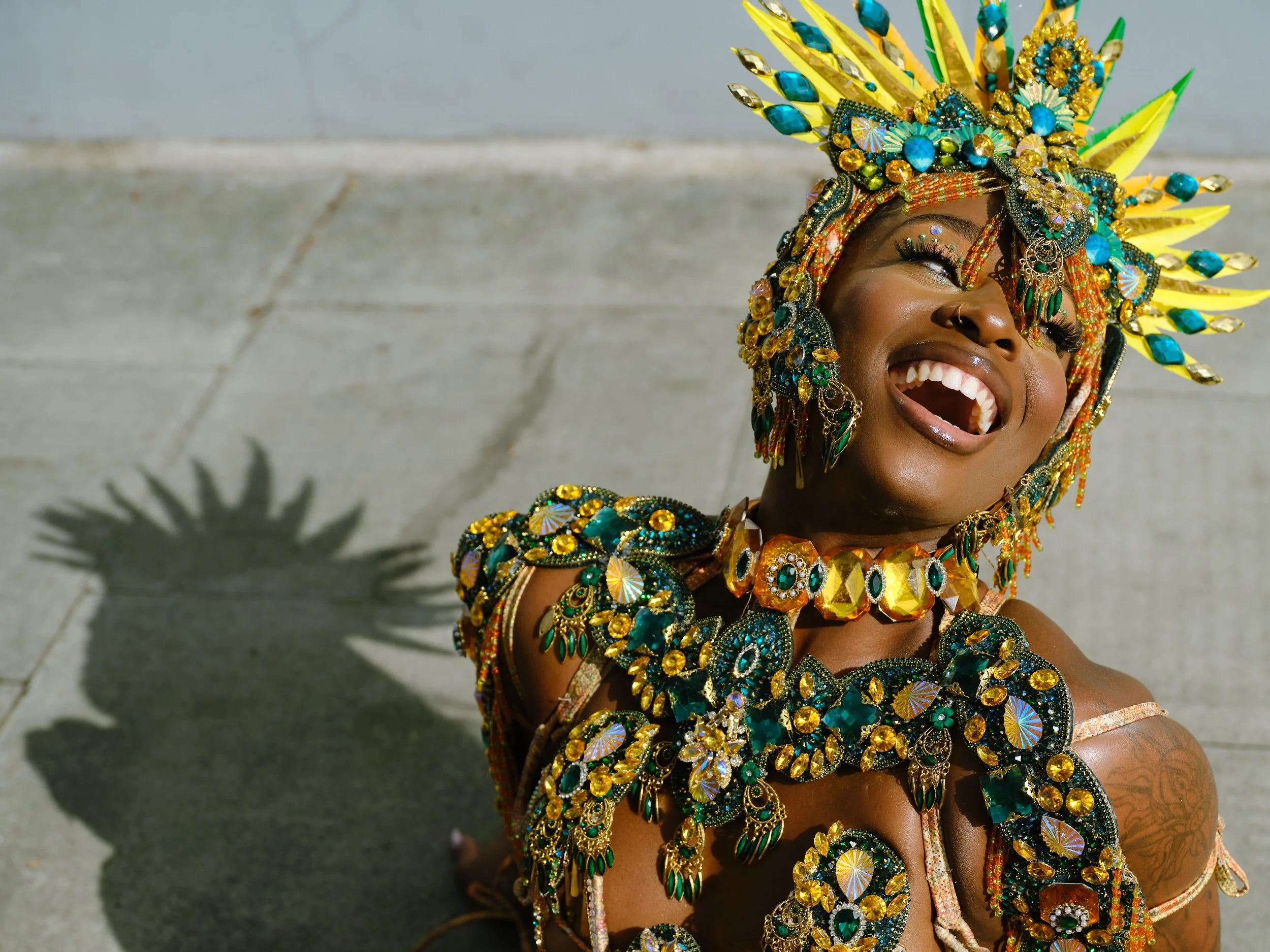 Woman dressed in a colorful, ornate costume with a large headdress decorated with yellow, blue, and green gemstones, smiling and looking upwards.