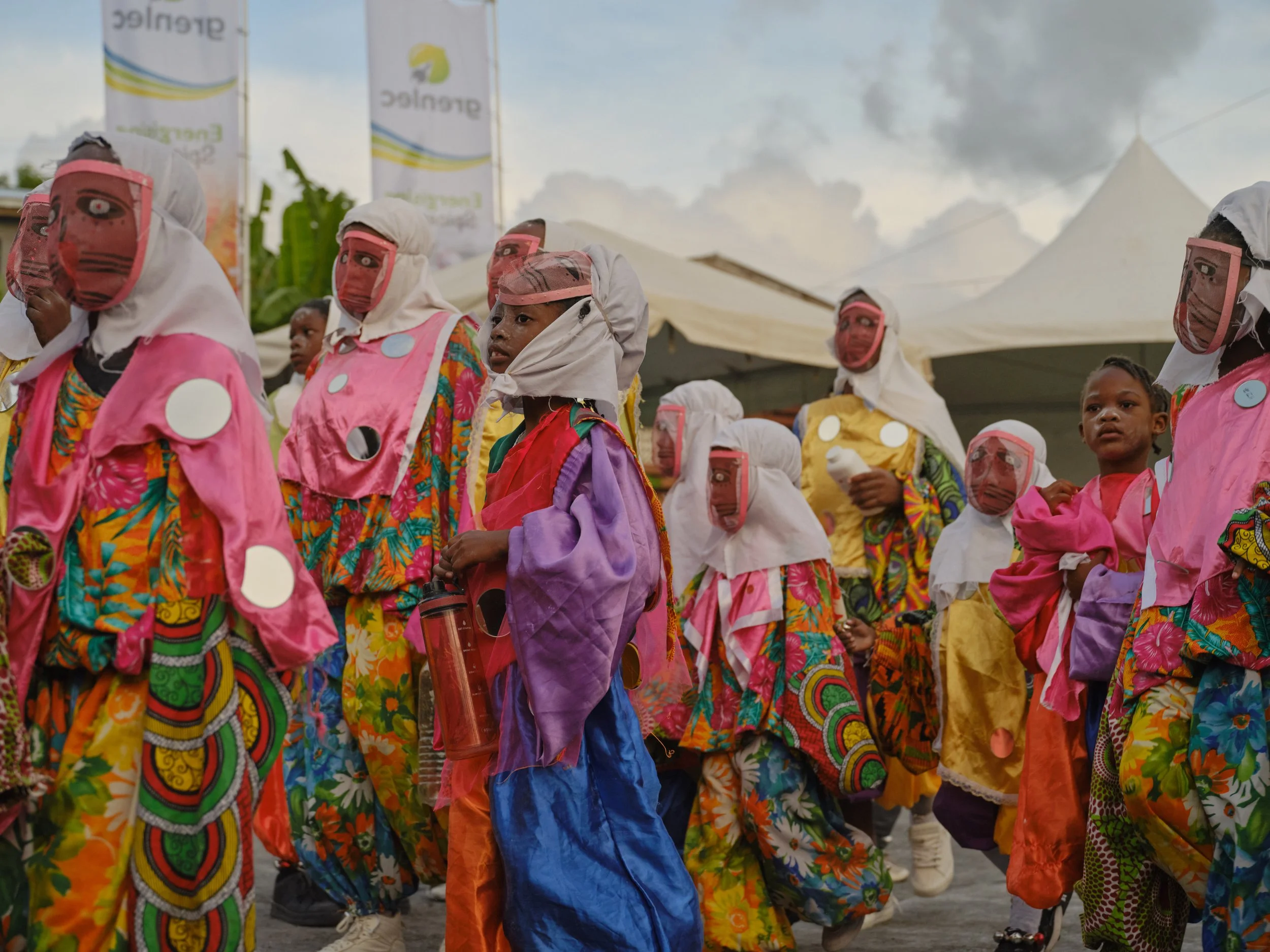 A large group of children dressed in colorful, traditional clothing, walking outdoors during daytime. Some wear face shields, and tents and banners are visible in the background.
