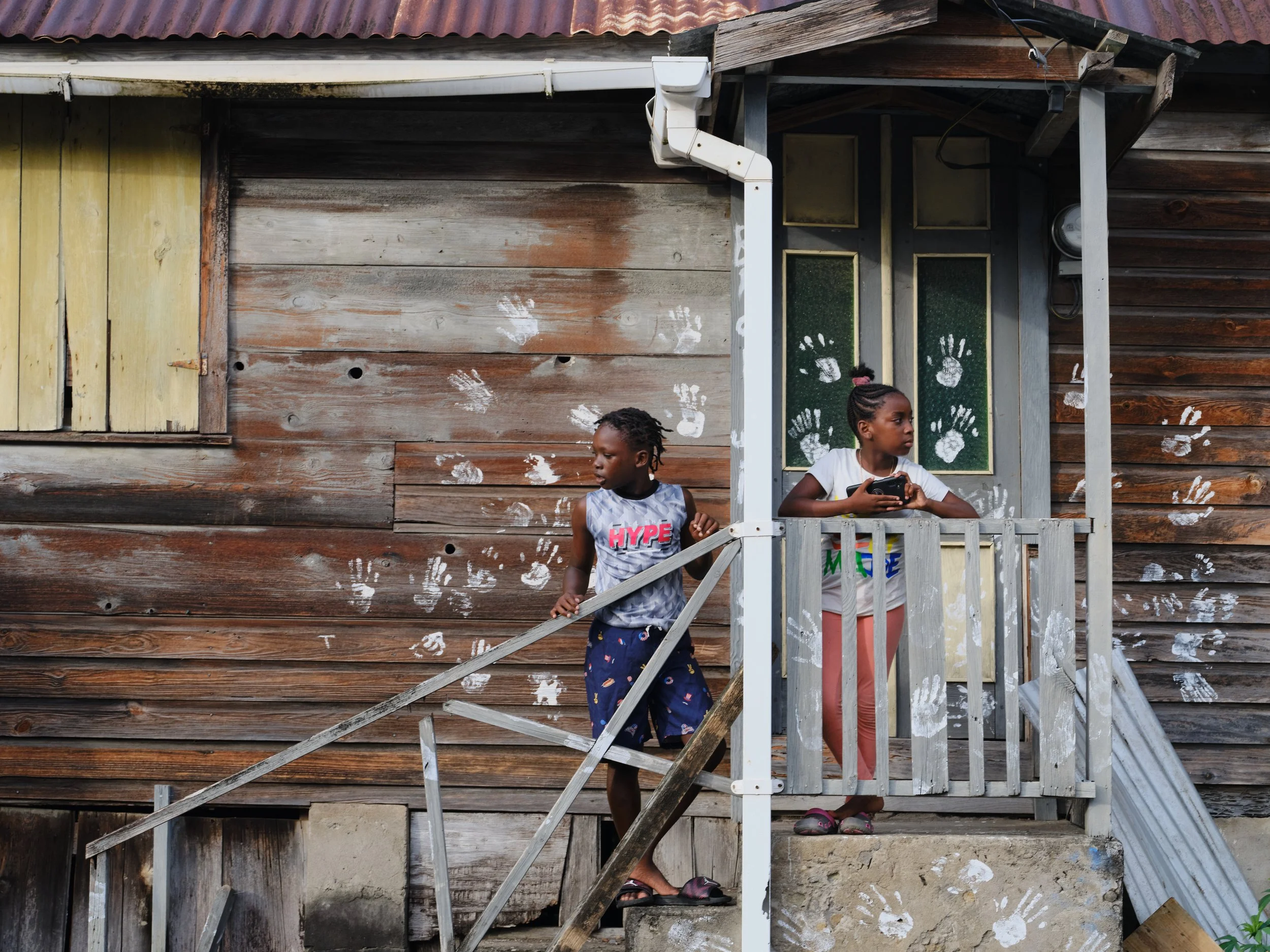 Two young girls standing on a porch with a weathered wooden wall behind them. The porch has a small roof and a white railing. Handprints are visible on the wall and windows. One girl is wearing a tank top with the word 'HYPE' on it, and the other gir