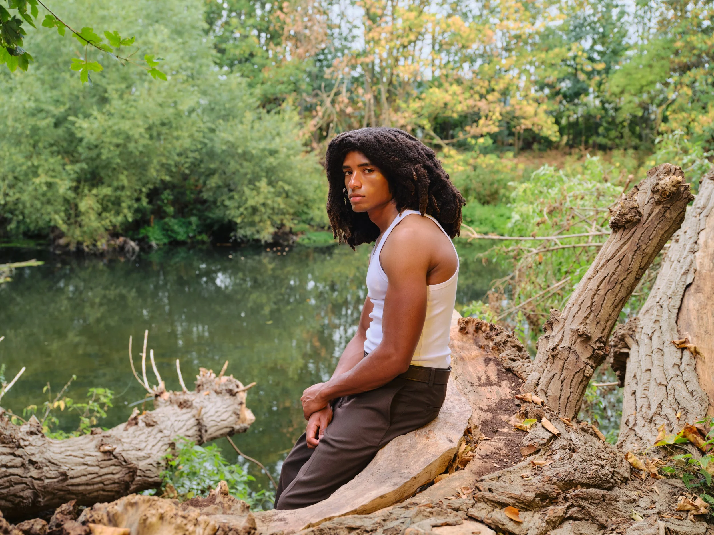 A young man with long dreadlocks wearing a white tank top and brown pants, sitting on a fallen tree trunk by a river in a lush green forest.