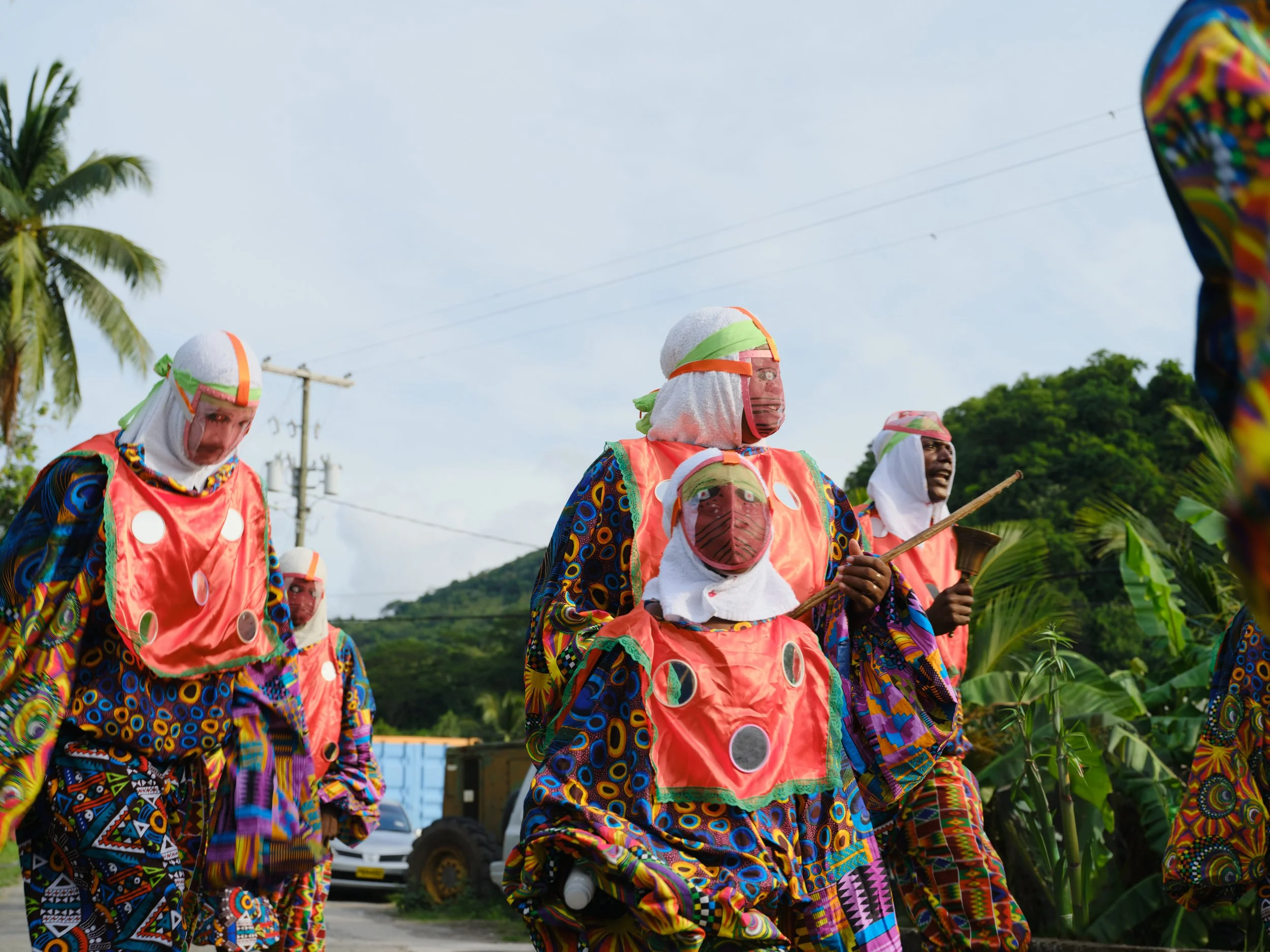 Group of people dressed in colorful traditional attire participating in a cultural event outdoors, with trees and utility poles in the background.