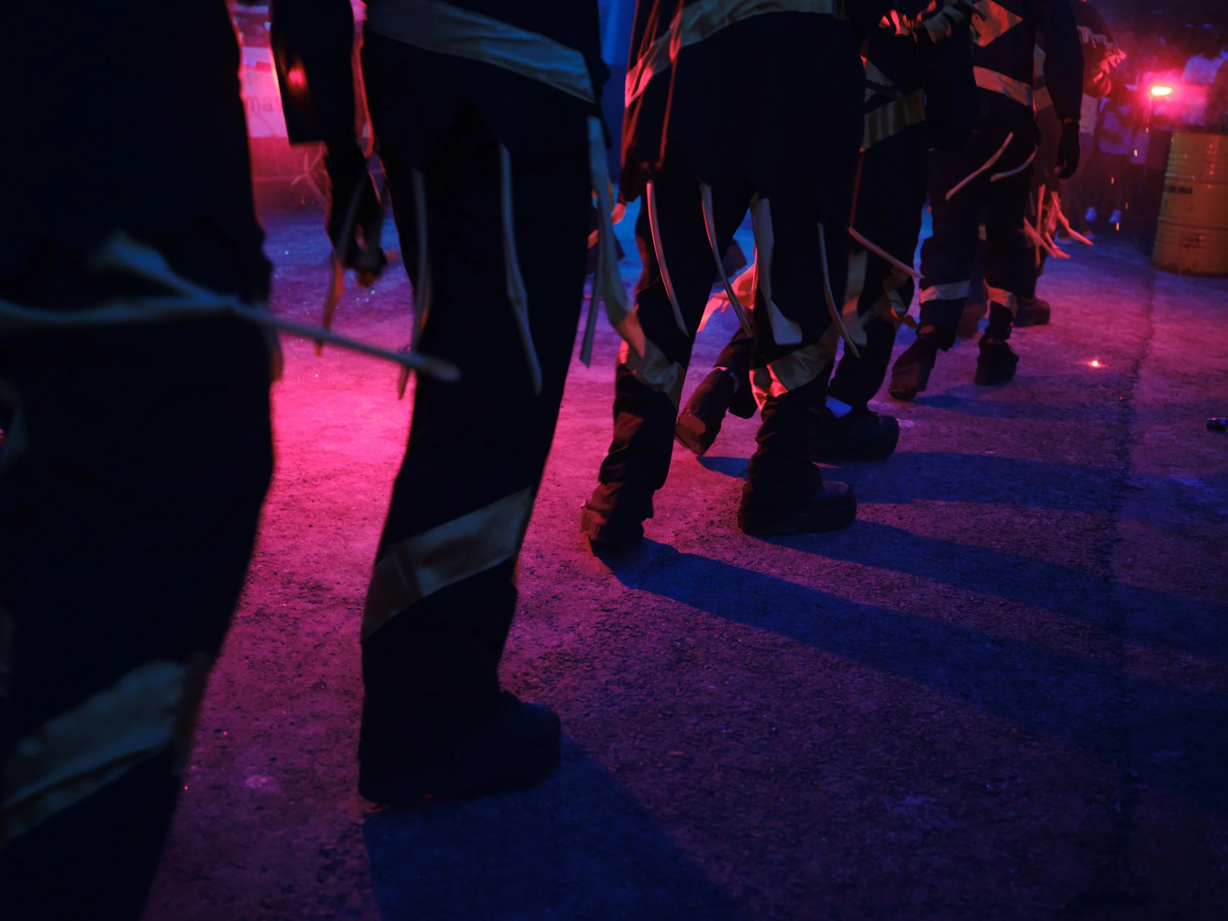 Line of workers dressed in dark uniforms with reflective stripes, standing in a row in an environment illuminated by red and blue neon lights.