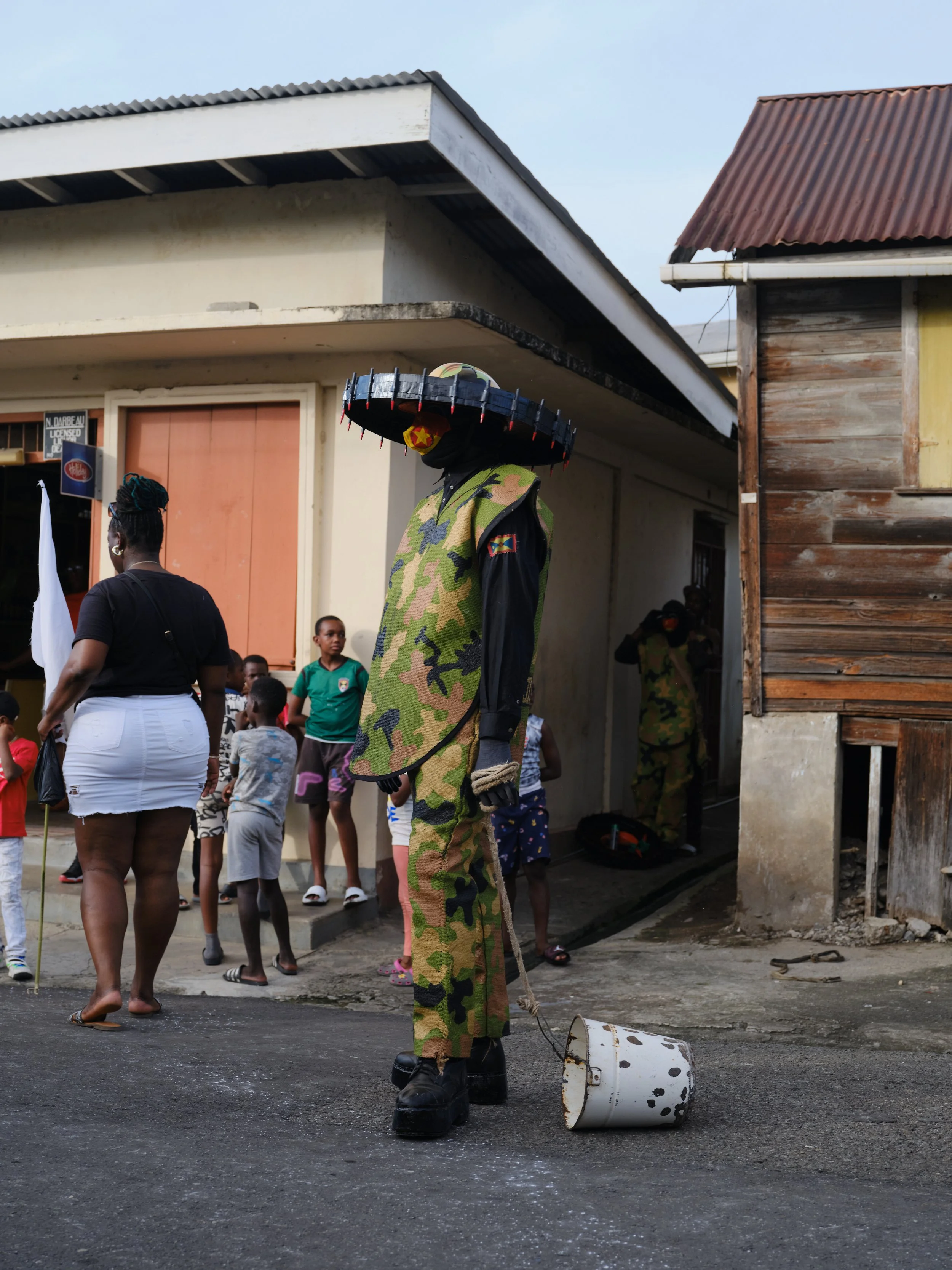 A person dressed in a camouflage outfit and oversized platform boots stands on a street next to a tipped-over metal bucket. In the background, there are people, mostly children, gathered near a building with a wooden exterior and a closed shutter, wi