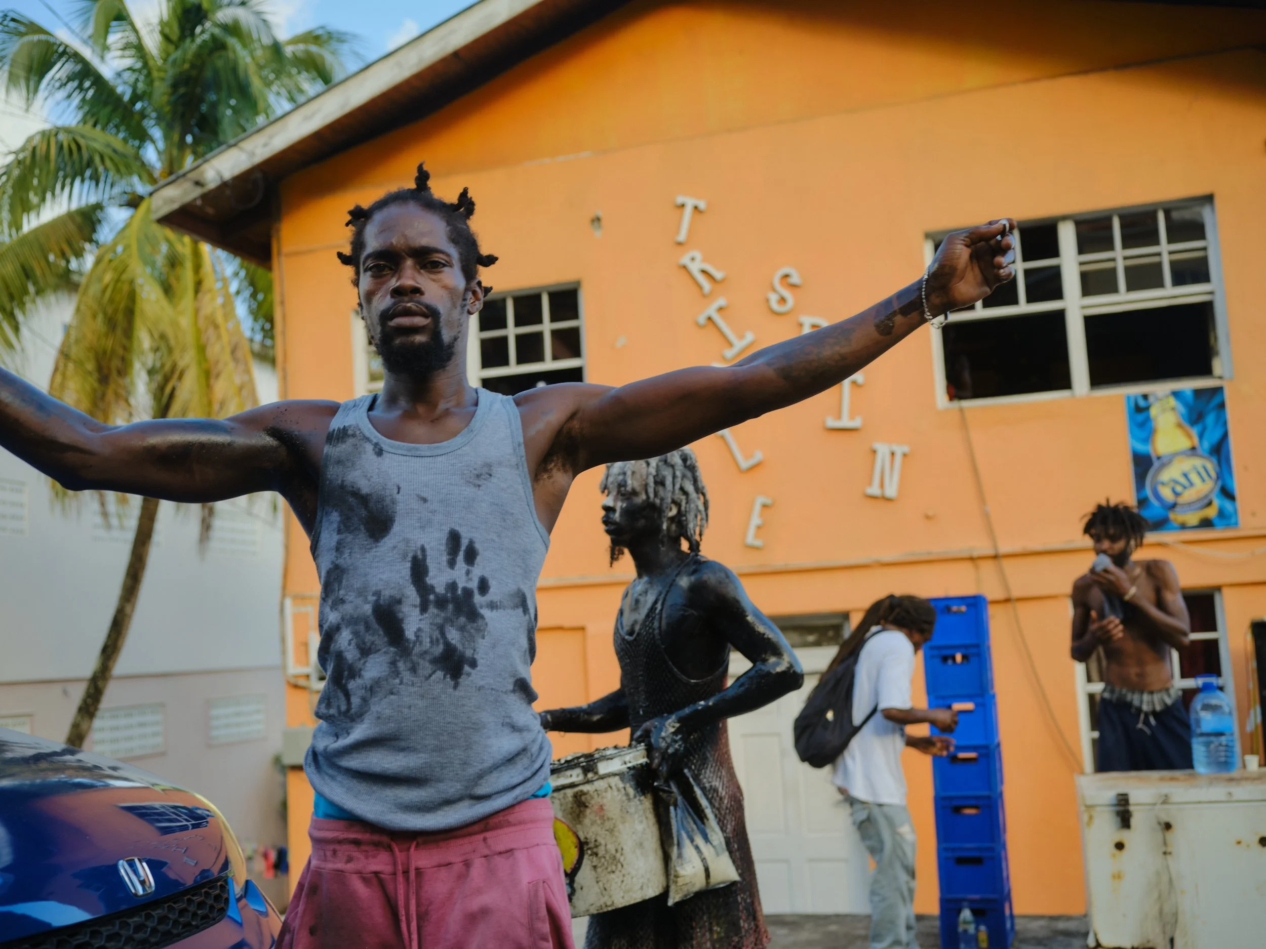 A man with dyed hair styled in bantu knots in front of an orange building with white letters spelling 'TRISTAN' on the wall, palm trees, and other people in the background.
