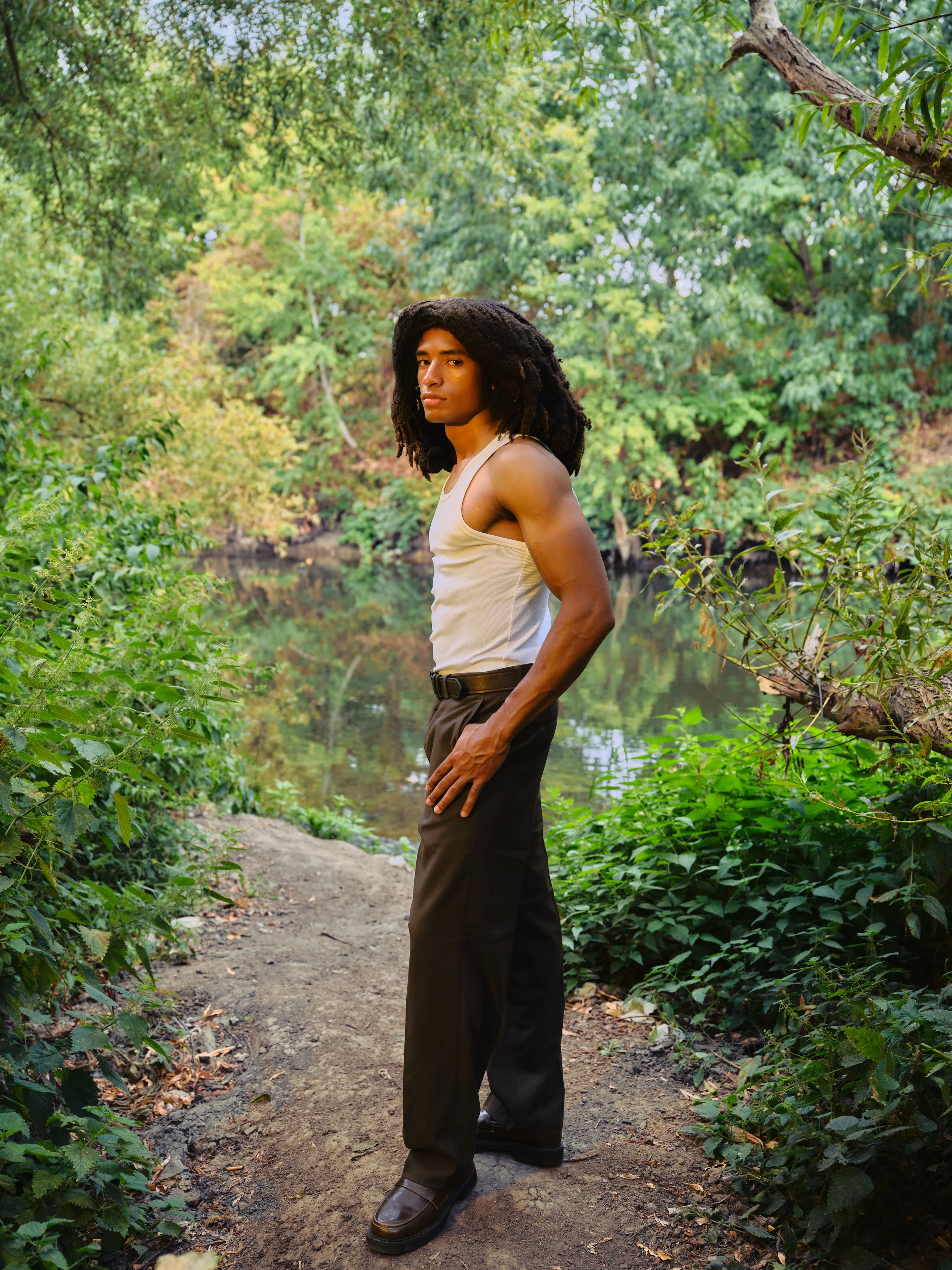 A person standing on a dirt trail in a lush green forest, looking at the camera with a serious expression, wearing a beige tank top, dark trousers, and brown shoes.