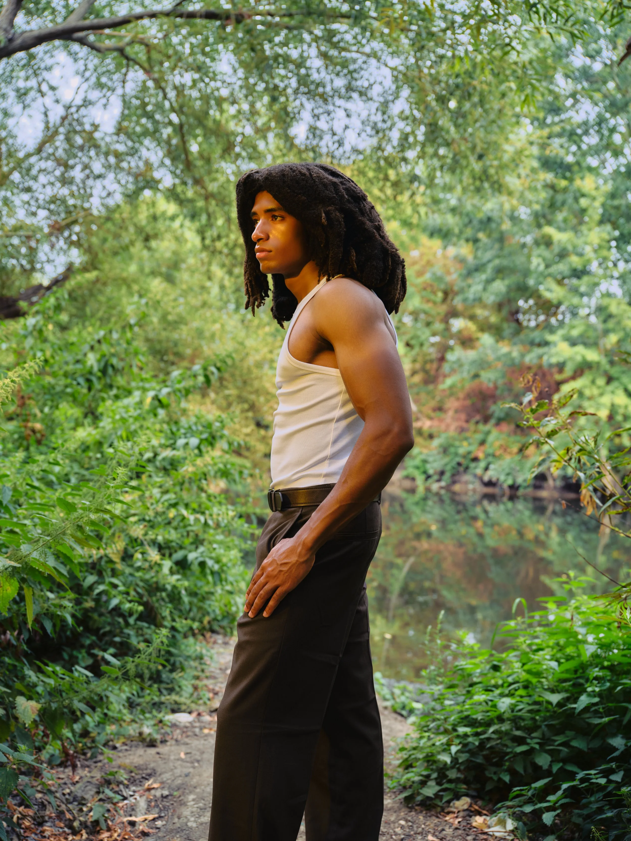 A person with long dreadlocks wearing a beige tank top and black pants standing on a forest trail surrounded by green foliage and trees.