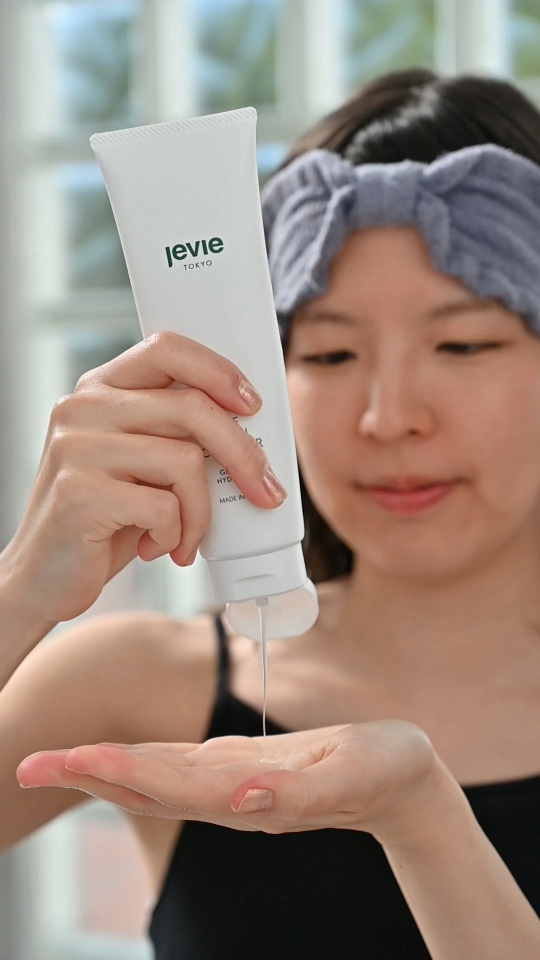 A woman with a gray headband and black tank top applying liquid from a white tube labeled 'levie TOKYO' onto her hand.