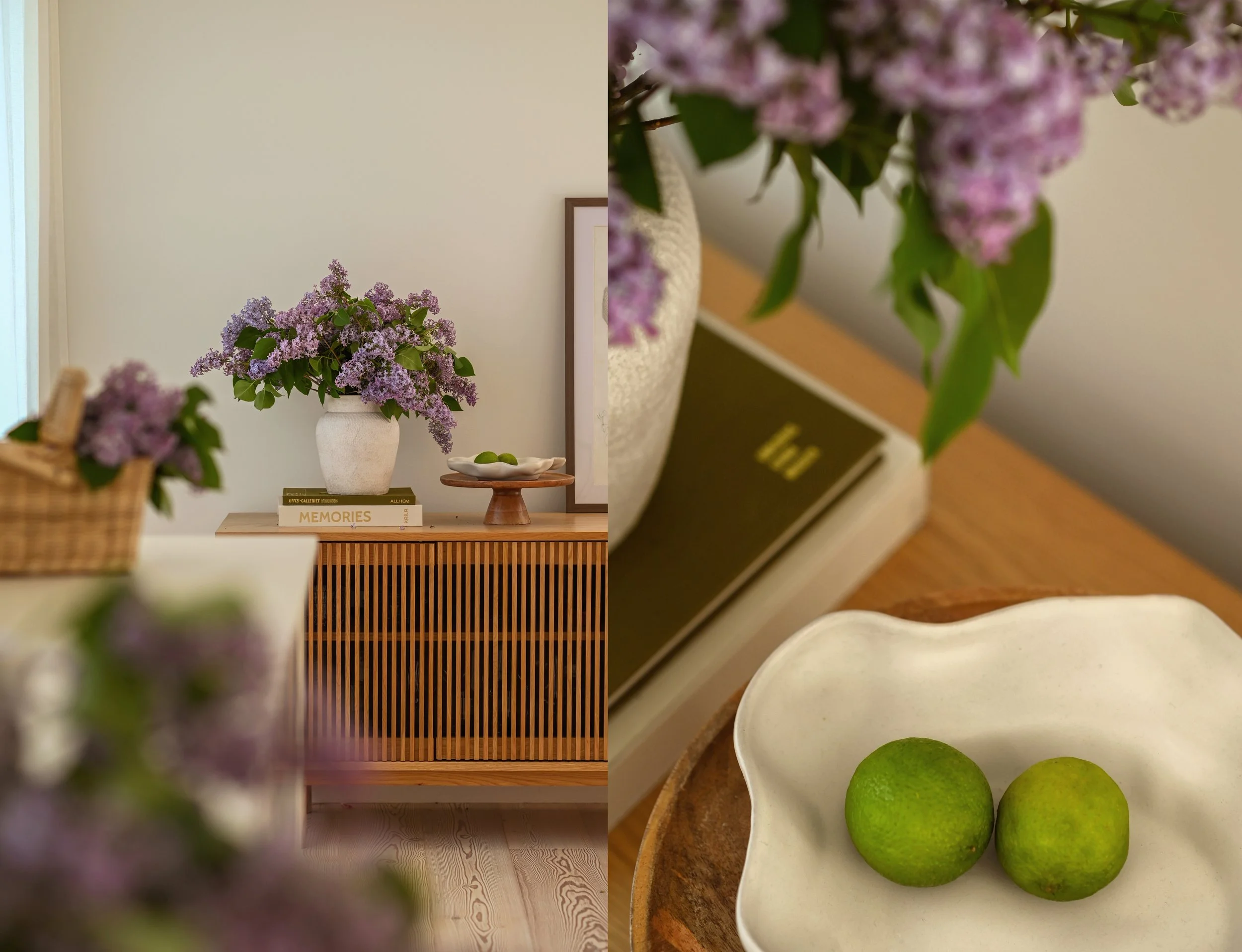 Close-up of purple lilac flowers in the foreground with a blurred indoor scene showing a vase of purple lilacs on a wooden table, books, and a lime green lime on a white dish in the background.