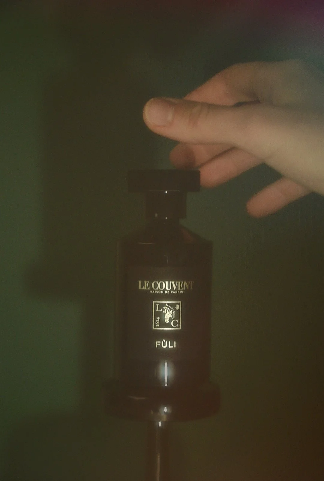 A person's hand holding a black perfume bottle labeled "LE COUVENT HAISON DE PARFUM" and "FÜLI" against a dark green background. Product photo. Branded content