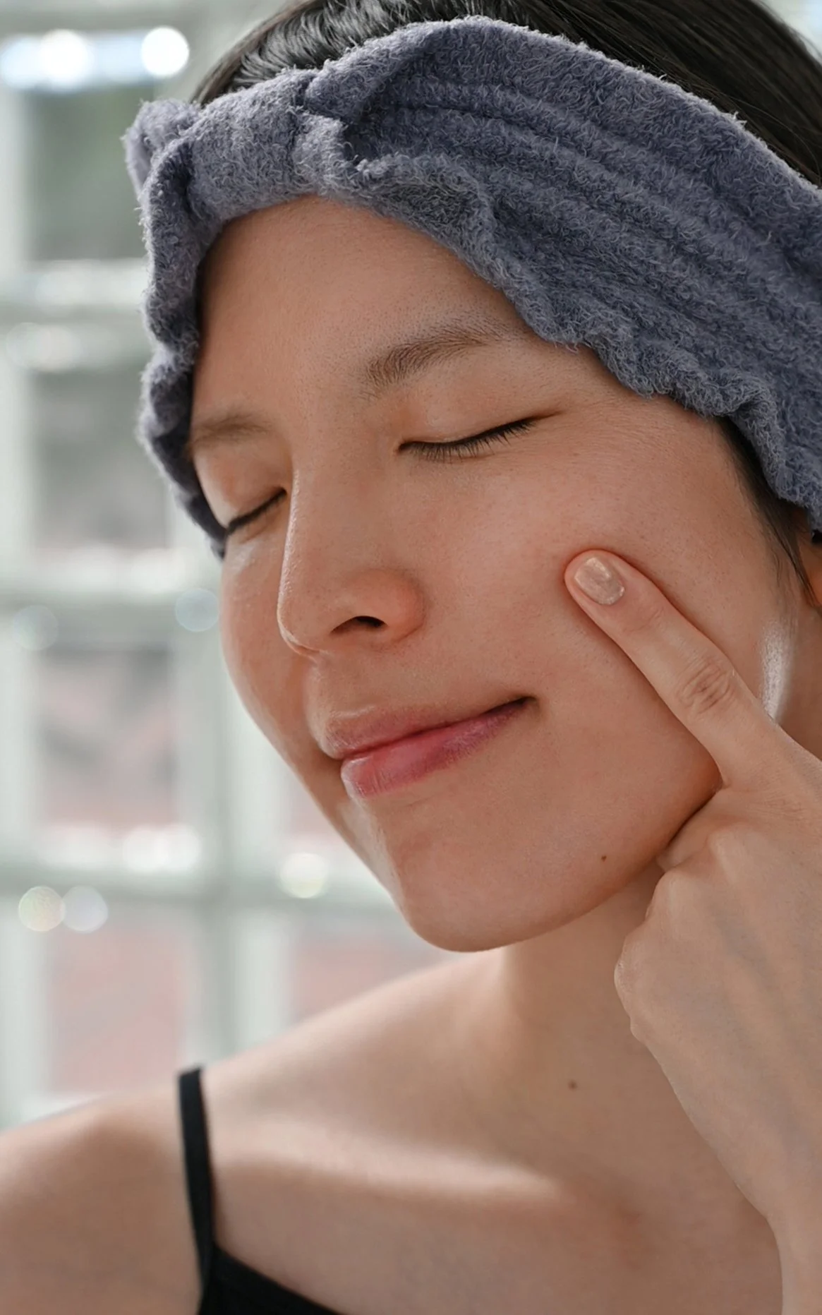 A woman with a towel wrapped around her head, gently pressing her finger on her cheek, with a relaxed expression, in a bright indoor setting.
