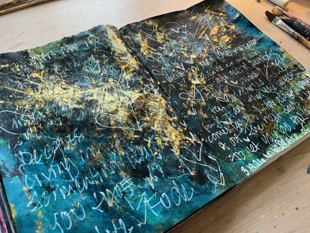 Open art journal spread with layers of teal and black paint, gold splatter, and handwritten words visible beneath the surface.