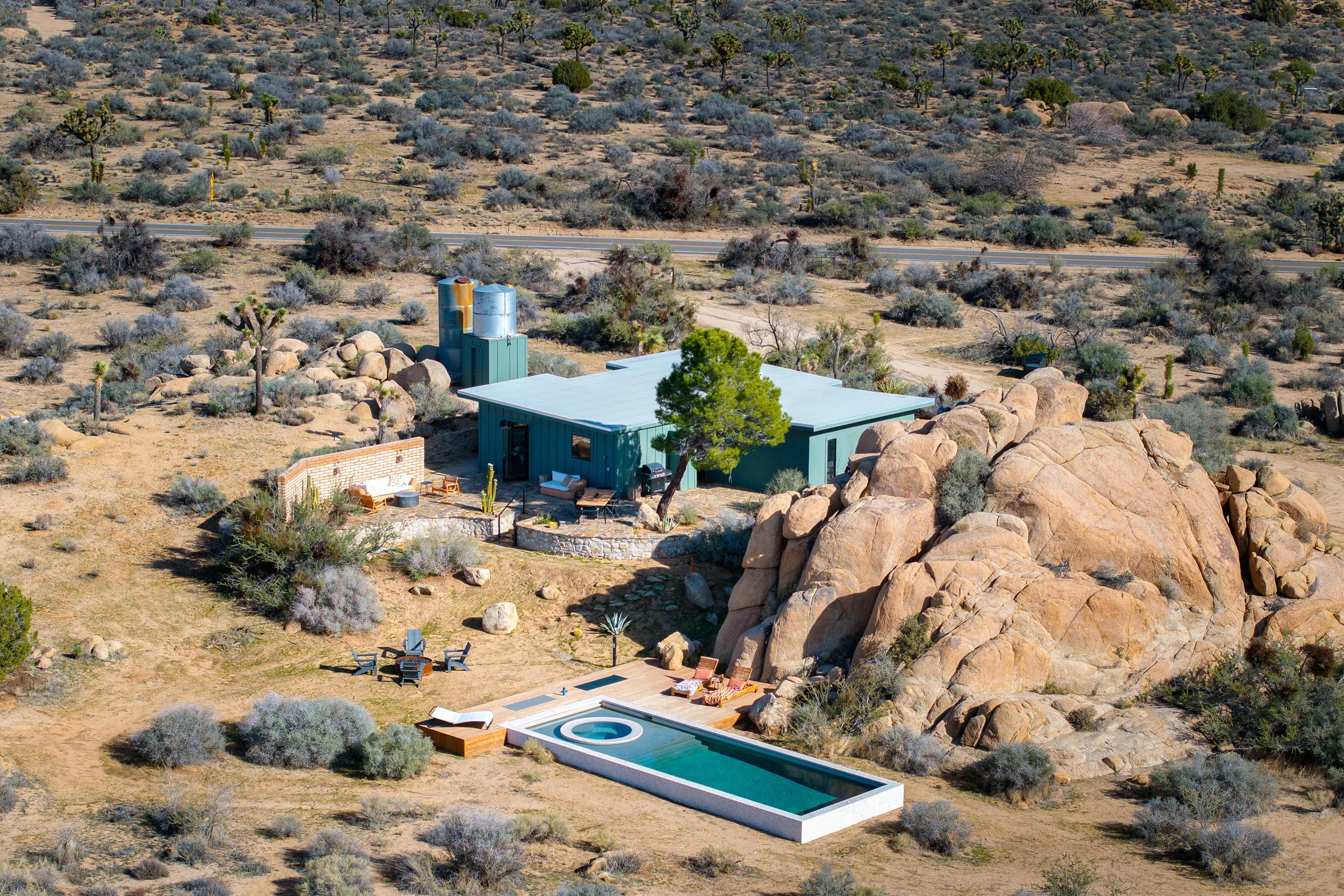 Aerial view of a desert landscape with sparse cacti and bushes, featuring a modern house with a swimming pool and a hot tub in the backyard.
