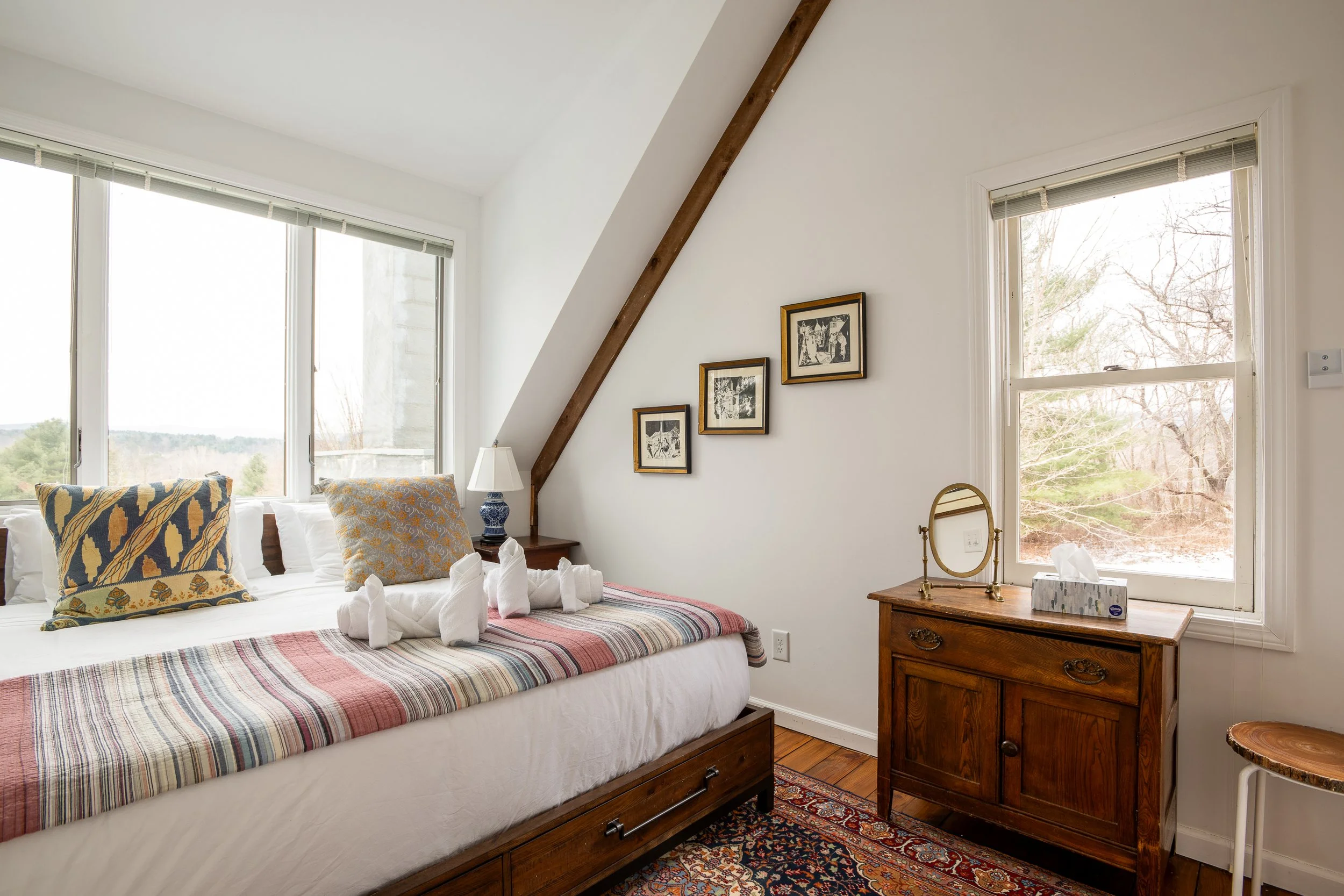 A cozy bedroom with two large windows showing a view of trees and sky. The bed has a white base with a multicolored striped blanket, decorated with pillows and towel swans. A wooden bedside table holds a blue and white lamp, a mirror, and tissue box, with a patterned rug underneath.