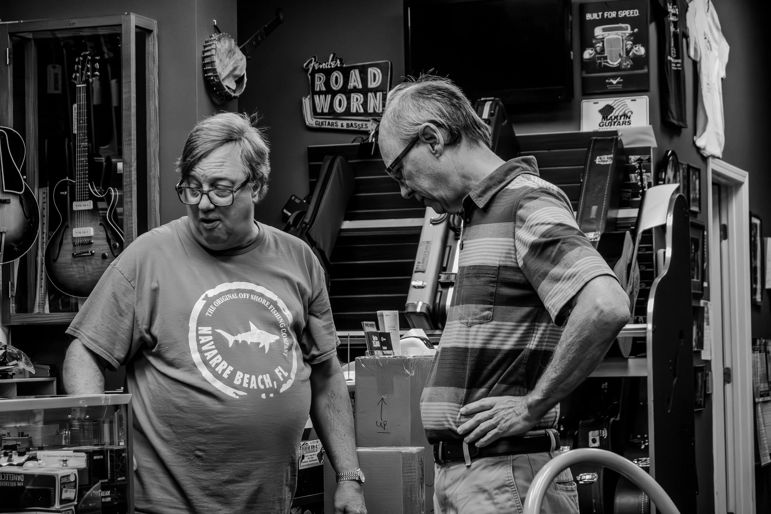 Two men in a music store looking at something on a counter. One is wearing a 'Navarre Beach, FL' t-shirt, the other a striped shirt. Guitars and musical equipment are visible in the background.
