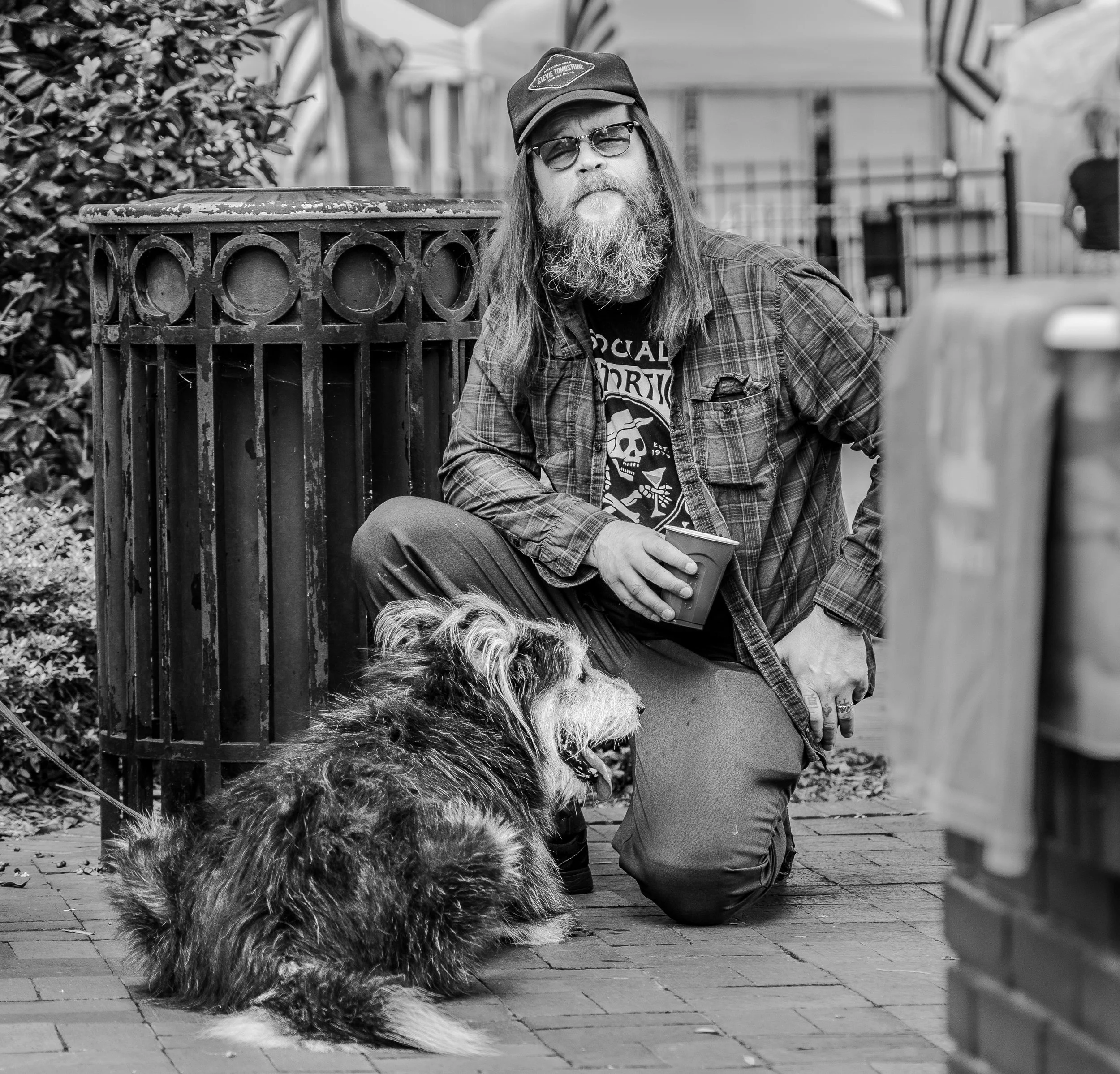 Man with long hair, beard, and sunglasses kneeling next to a sitting fluffy dog while holding a cup, in a black and white street scene.