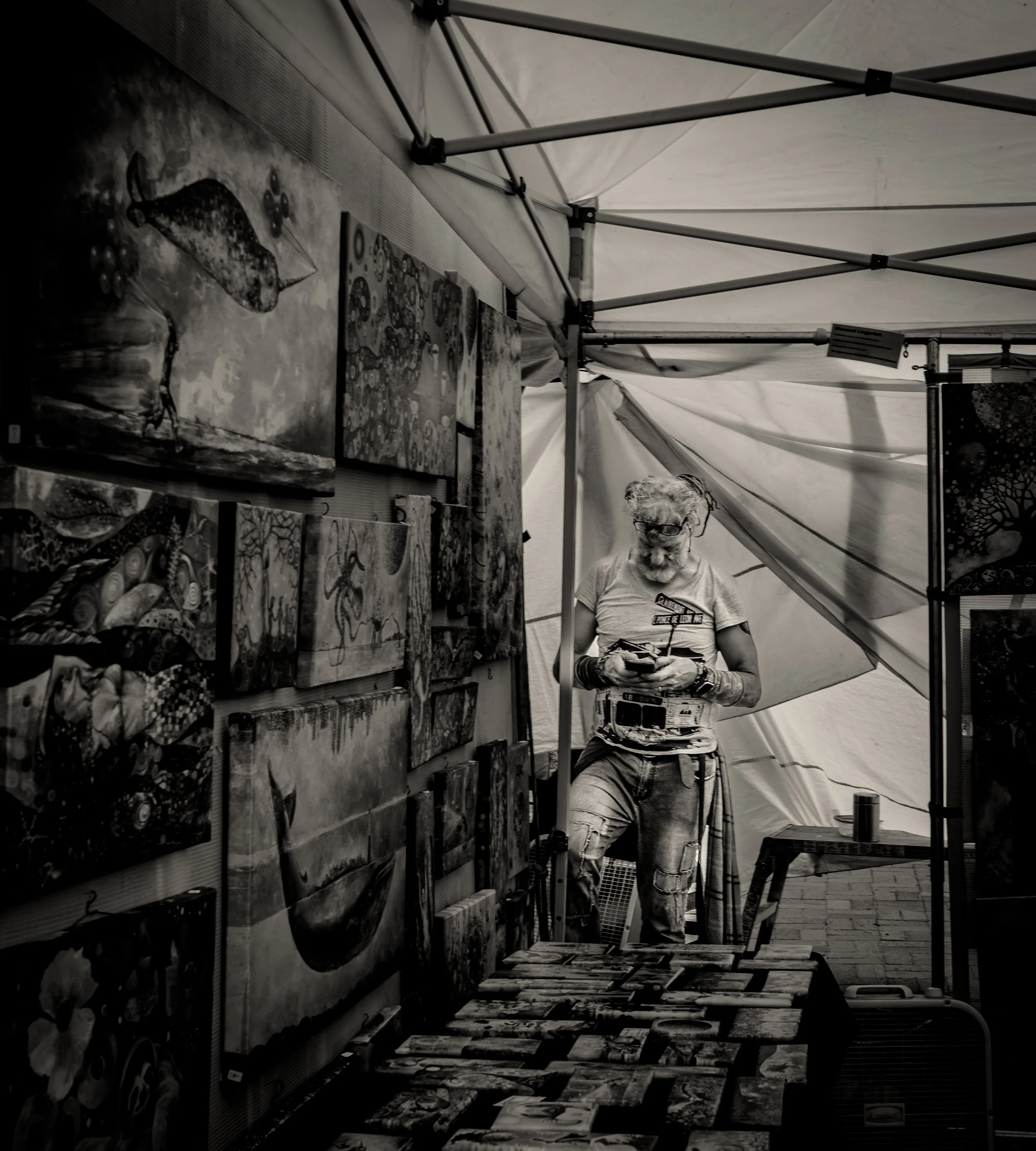 Black-and-white photo of an art exhibit in a tent. A person examines art on display, surrounded by various paintings with abstract and surreal designs.
