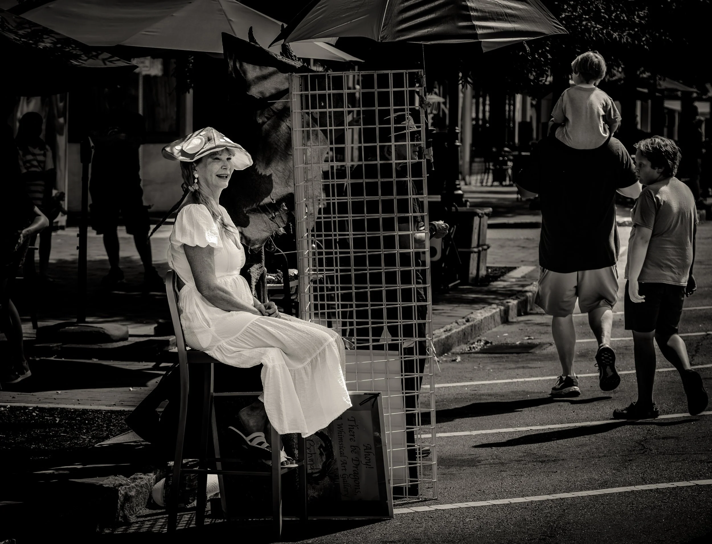 Black and white image of a woman in a white dress and hat sitting on a chair at an outdoor market or fair, with people walking by in the background.