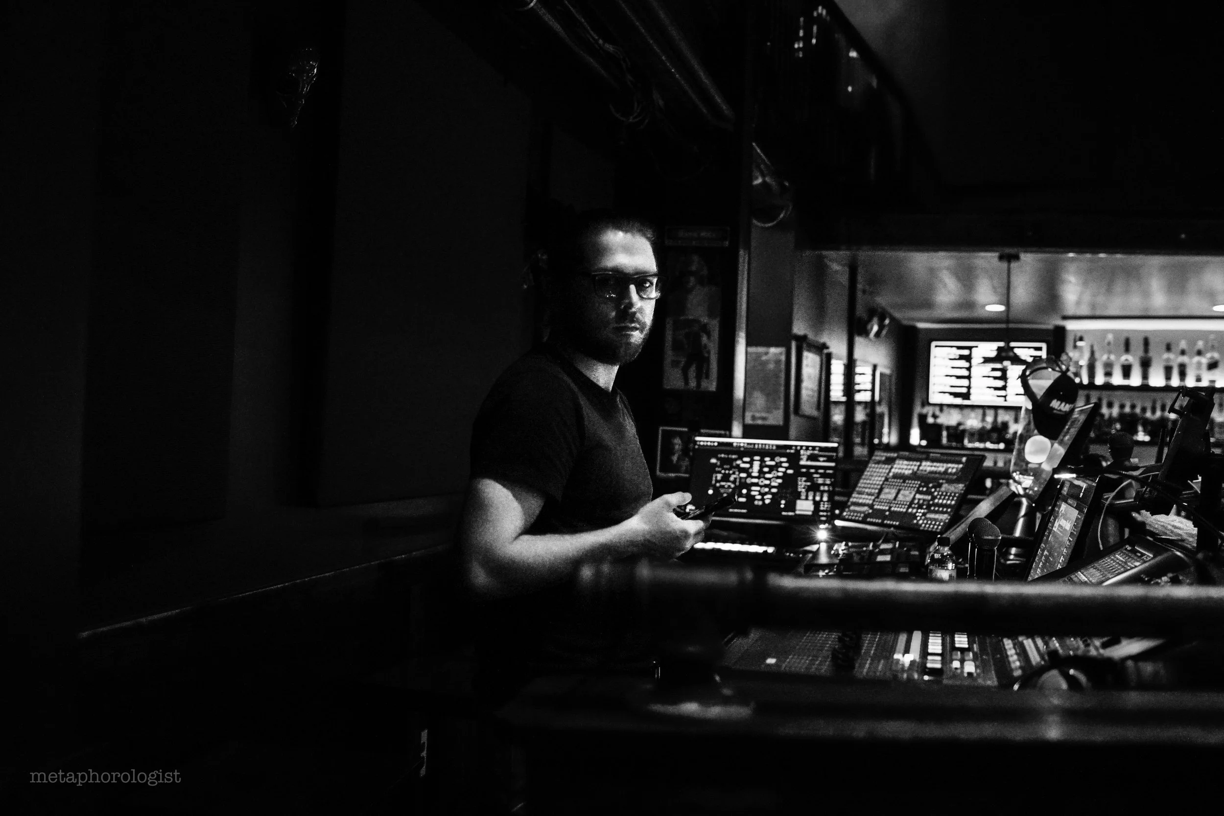 Black and white photo of a man in a dark room with audio equipment and multiple screens, possibly a sound engineer, holding a phone.