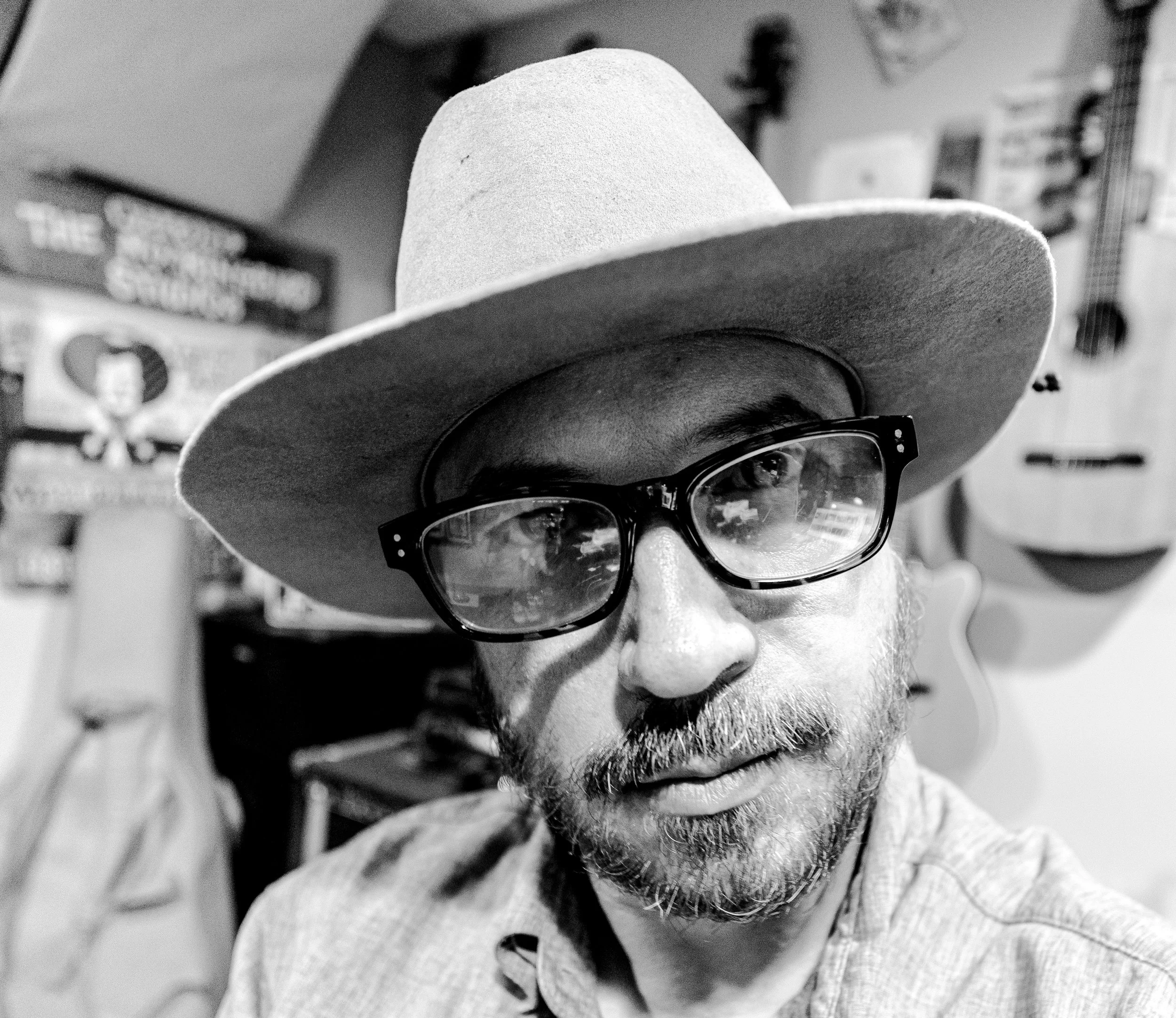 Black and white portrait of a man wearing glasses and a hat, with guitars and posters in the background.