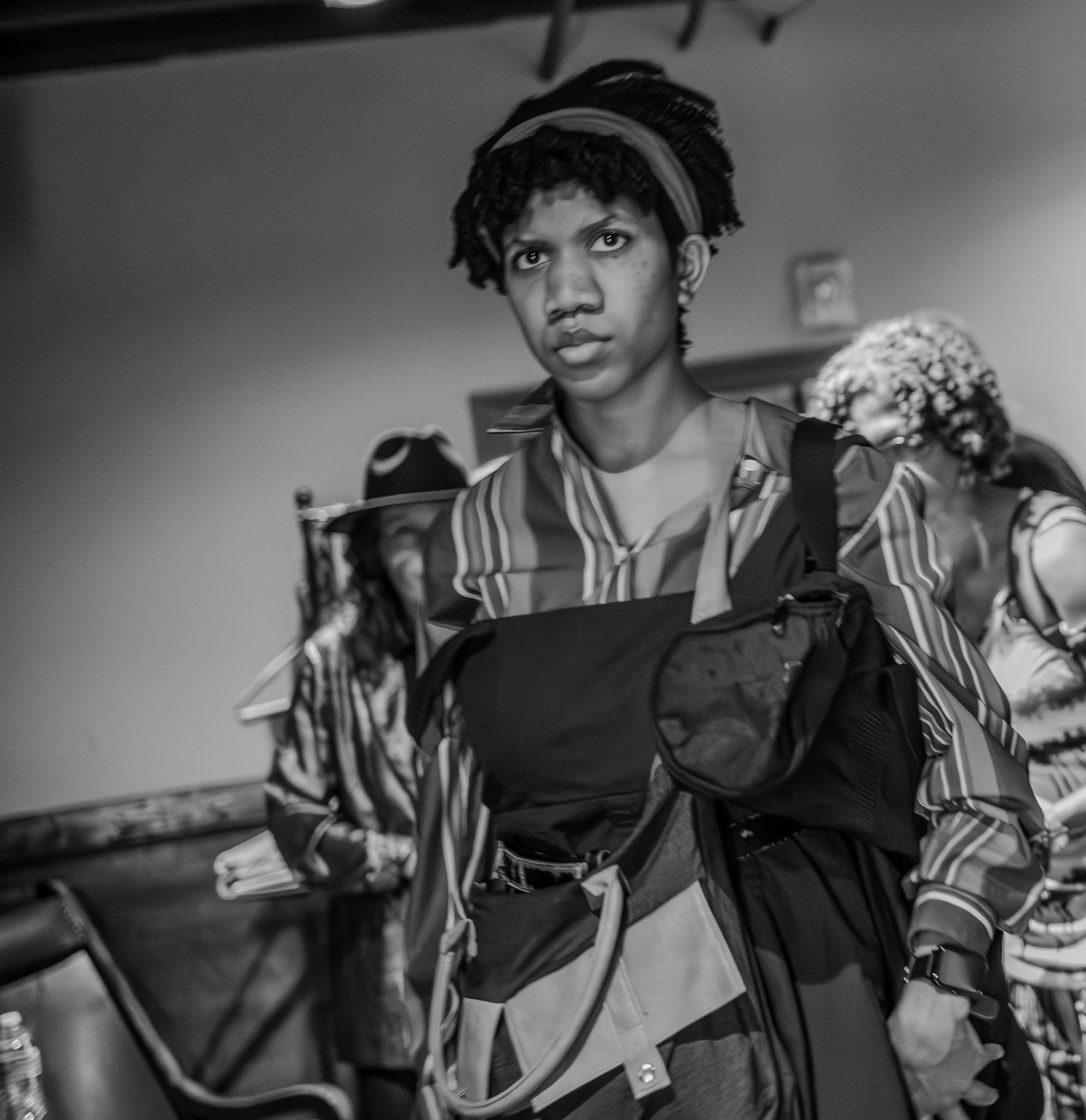 Black and white photo of a person in a striped shirt carrying multiple bags, with others in the background.