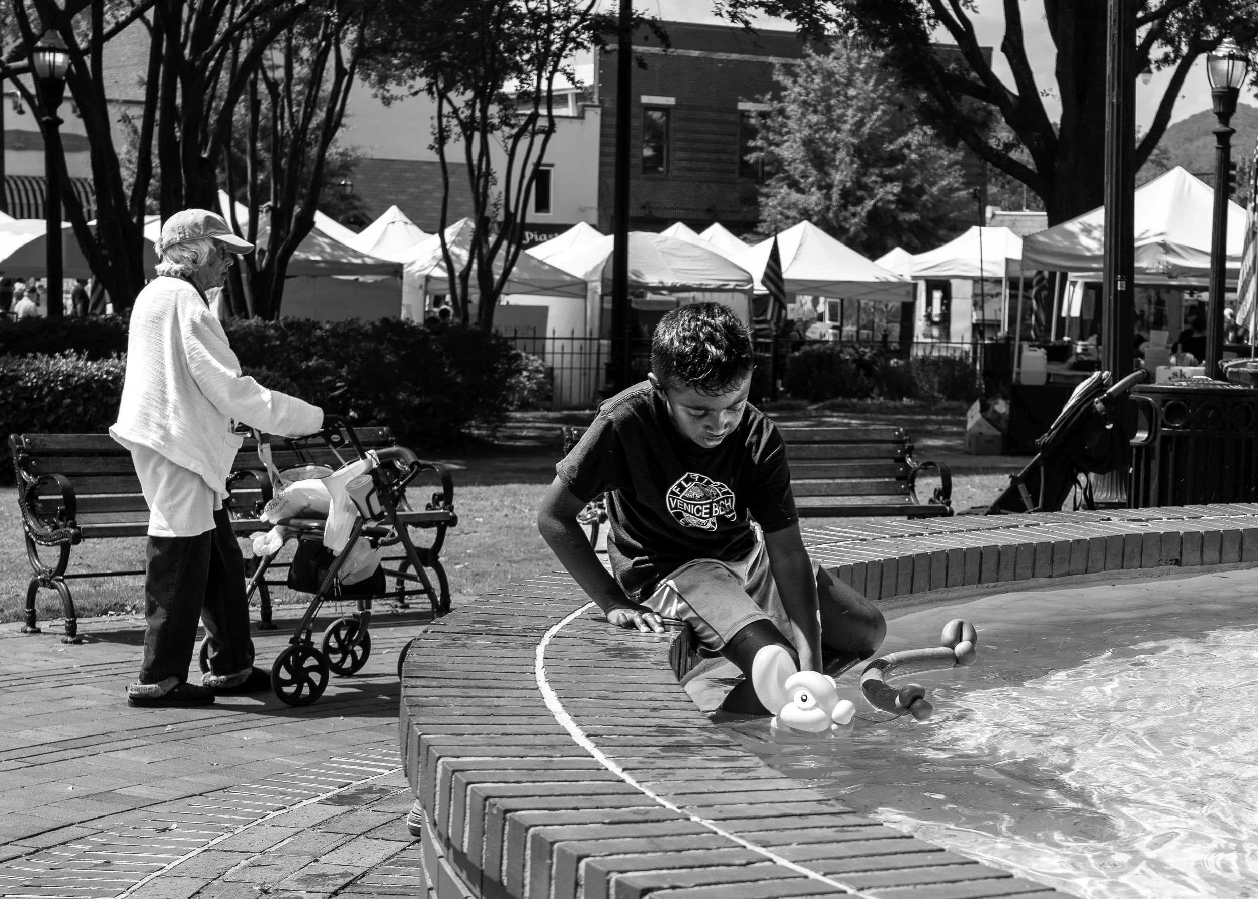 Black and white photo of a boy sitting on the edge of a fountain playing with a toy duck in water, with background of an elderly person walking with a walker and market tents visible.