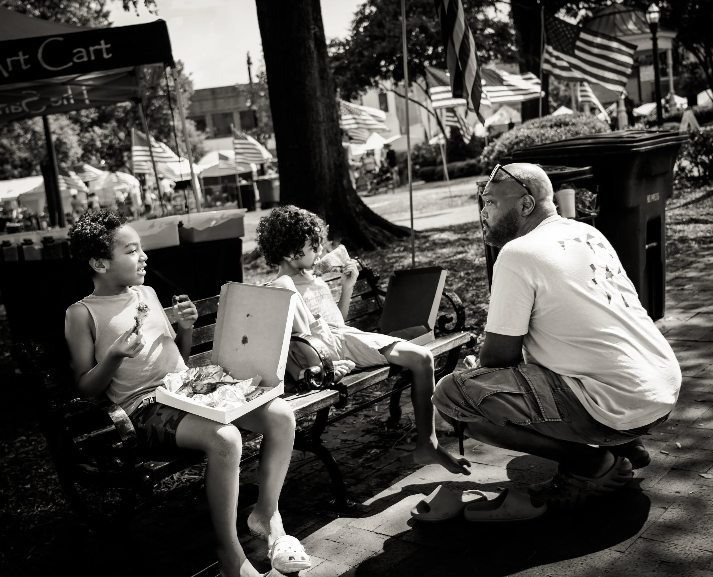 Black and white photo of two children sitting on a bench eating food from a pizza box, with a man crouching nearby. American flags and a tent can be seen in the background, suggesting a park or outdoor event.