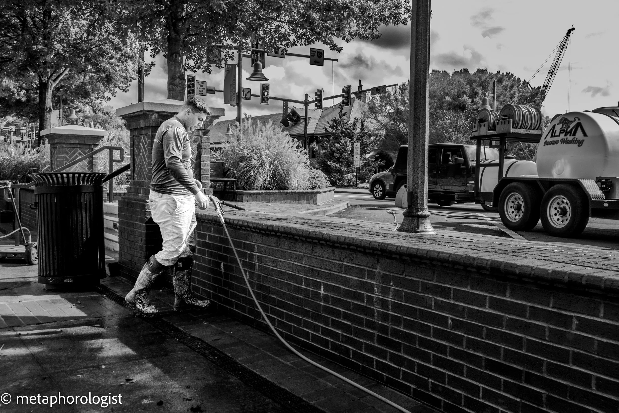 A person pressure washing a brick wall on a city sidewalk. A truck with a pressure washing tank is parked nearby. The scene includes trees and traffic lights in the background. The photo is in black and white.