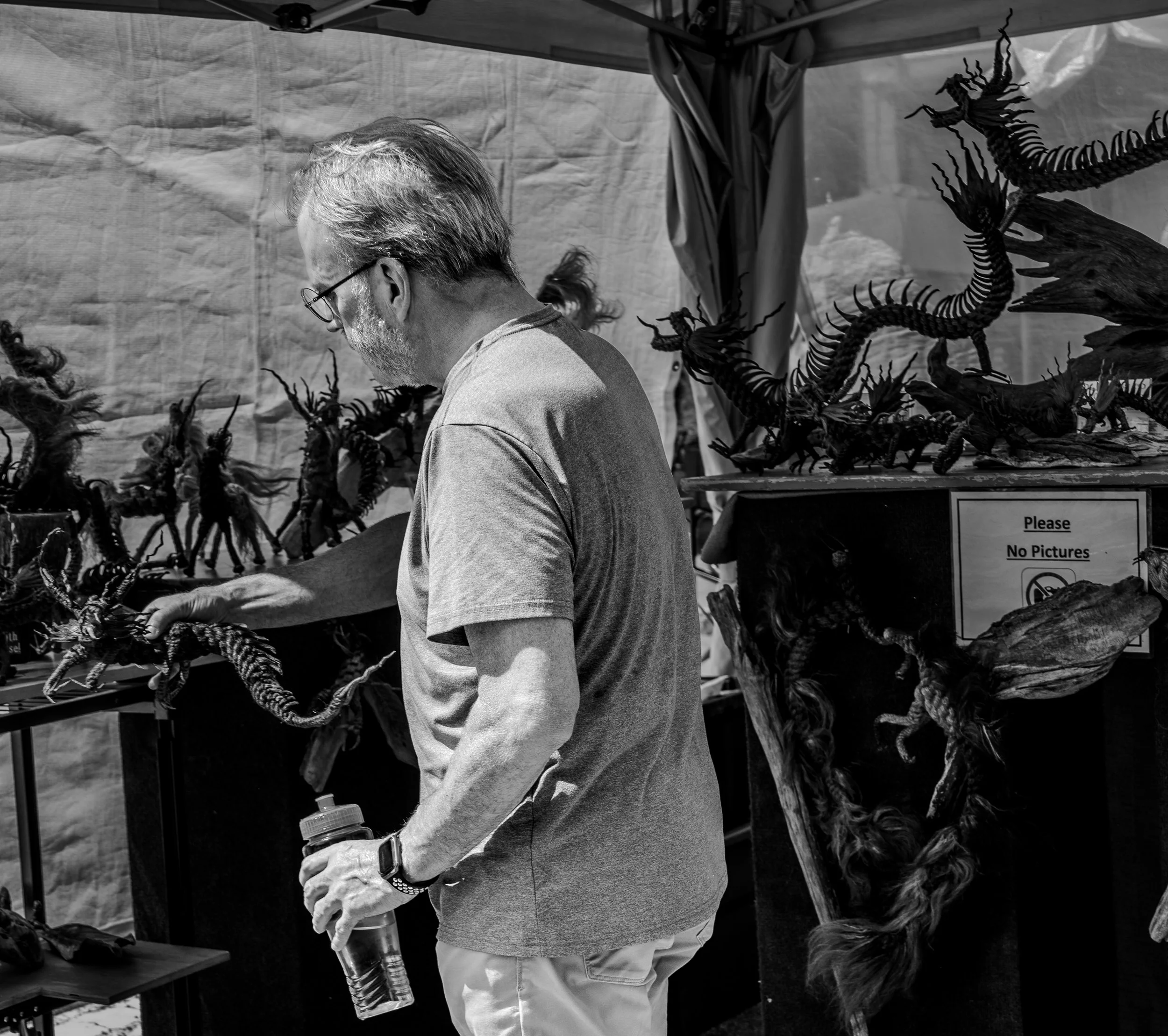 Black and white image of a man observing intricate sculptures on display at an outdoor art market. A "No Pictures" sign is visible.