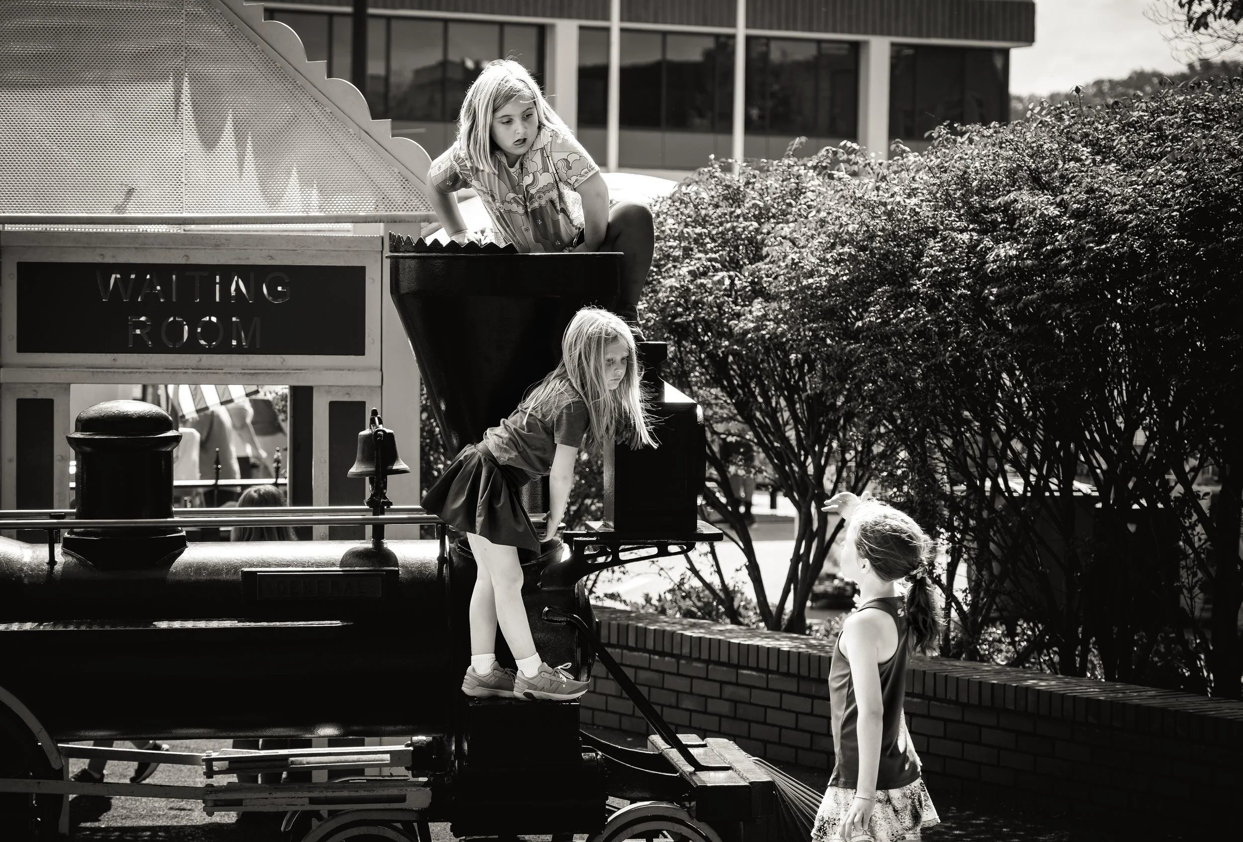 Three children playing on a vintage train display in front of a building with a "Waiting Room" sign.
