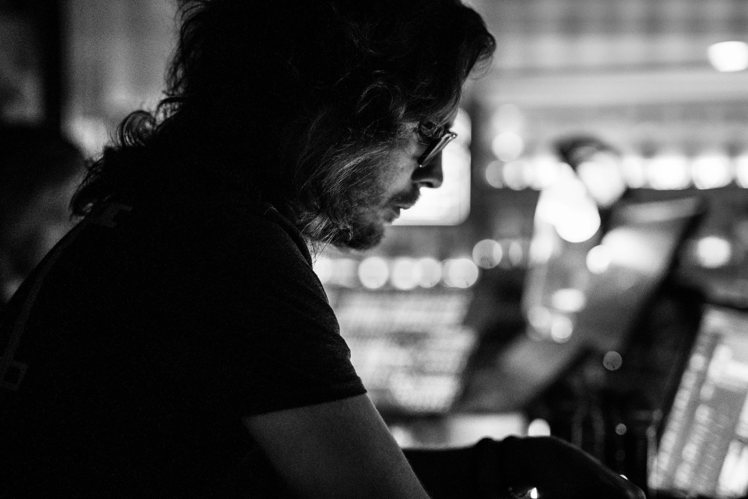 Black and white photo of a person with long hair and glasses in profile, focused on a computer screen with a blurred background.