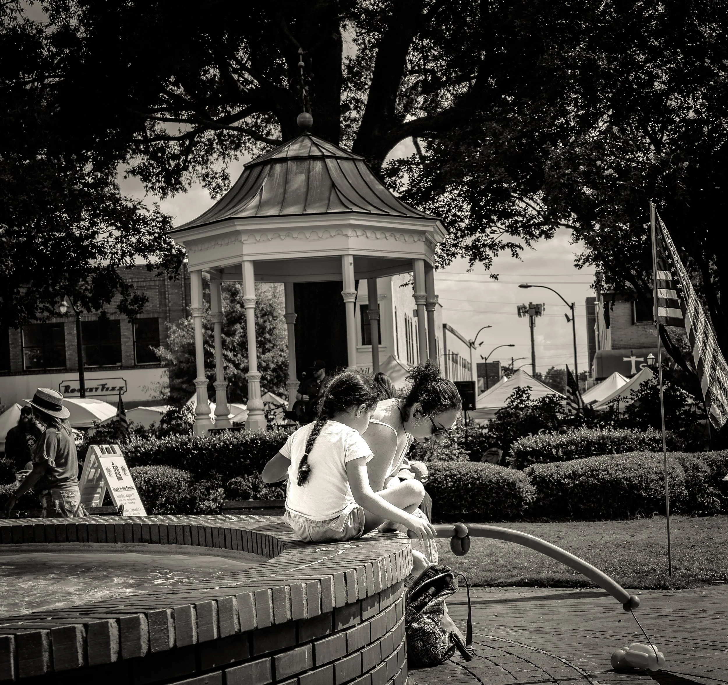 Black and white photo of a woman and child sitting by a fountain in a park, with a gazebo and American flag in the background.