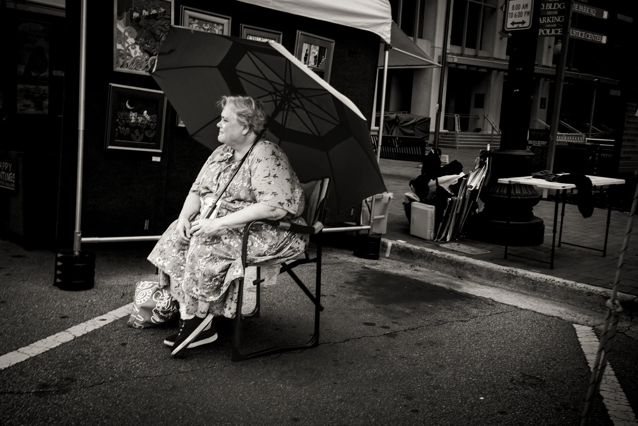 Woman sitting under an umbrella at an outdoor art exhibit with framed paintings behind her. Monochrome photograph.