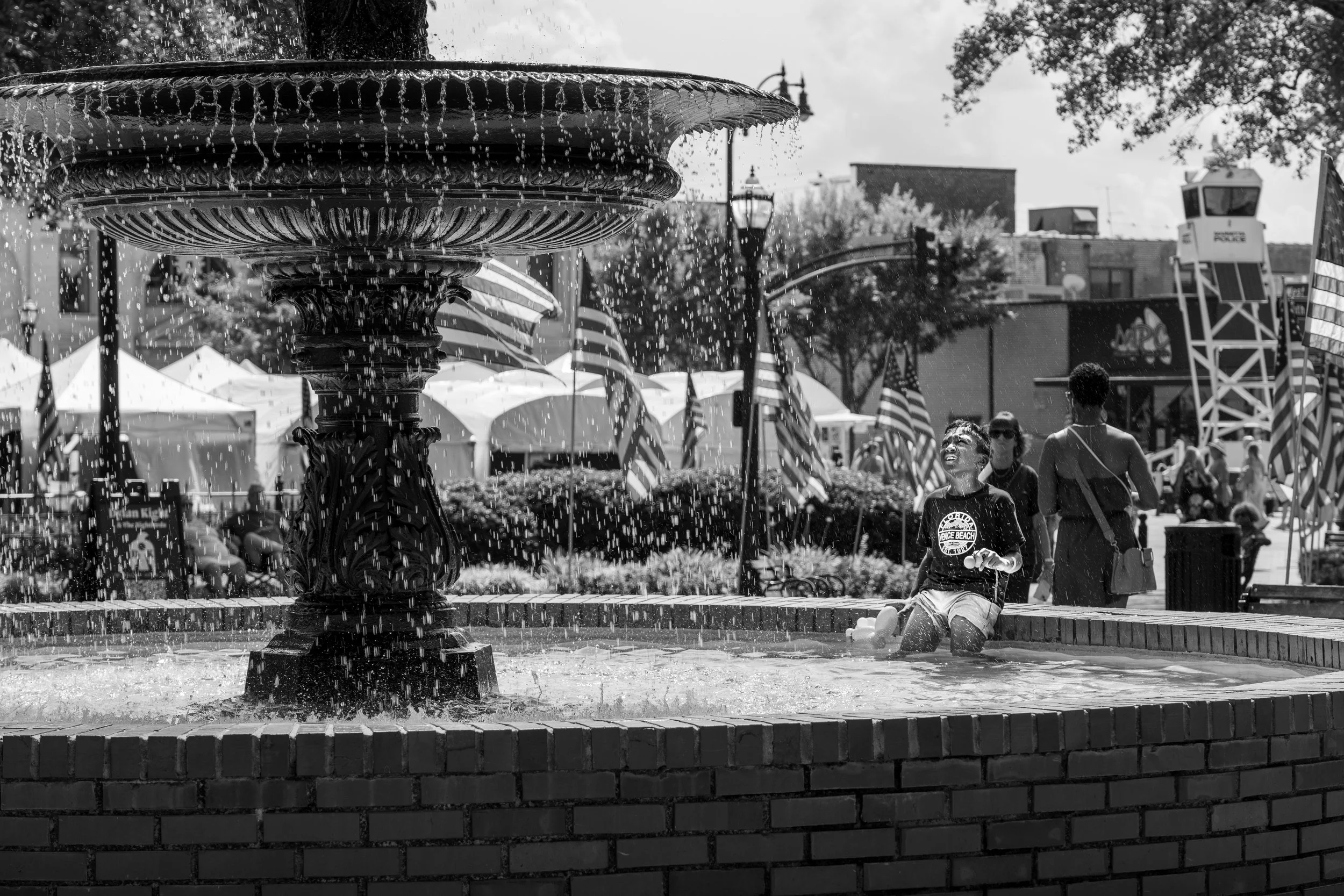 Black and white image of a fountain with American flags in the background, featuring a person sitting in the water laughing. Several tents and people are visible, suggesting a festive outdoor event.