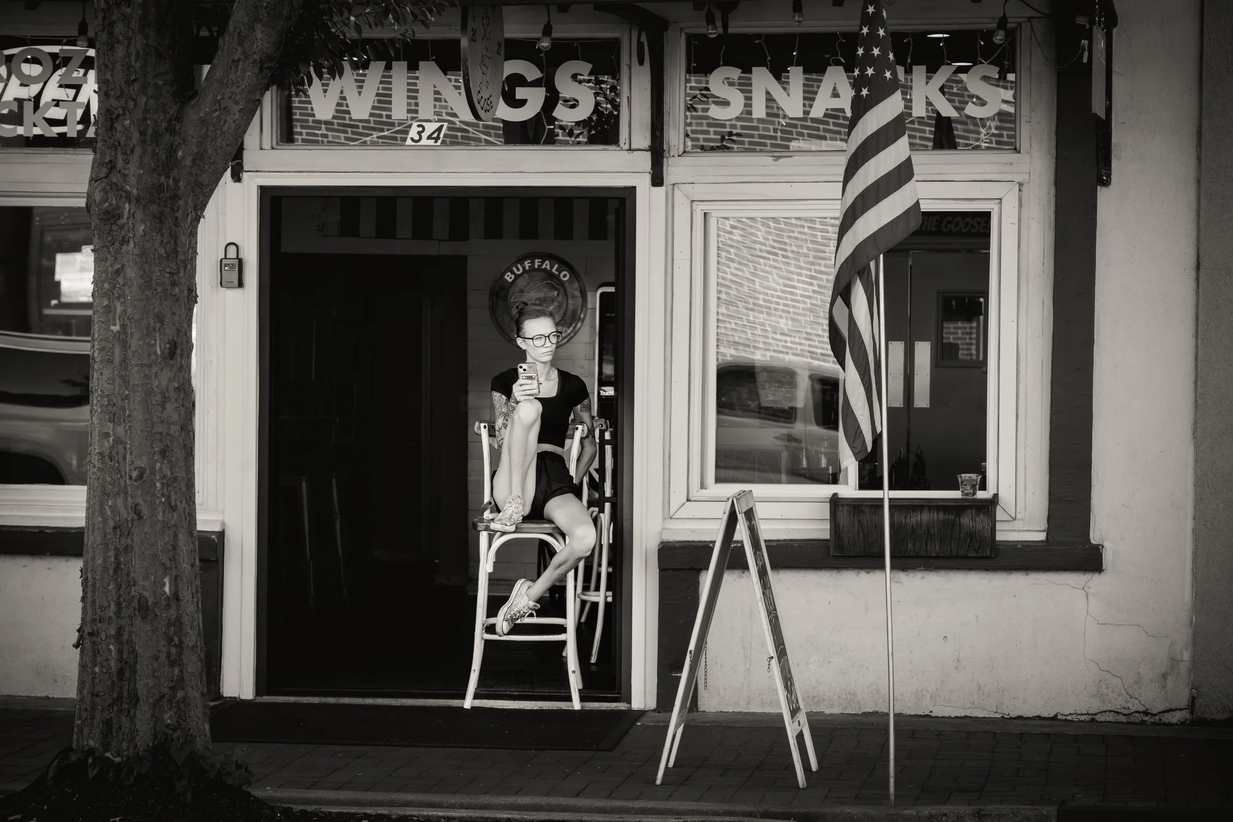 Black and white photo of a person sitting on a stool outside a storefront with "Wings Snaks" signage. The person is using a smartphone. An American flag and a sandwich board are nearby.