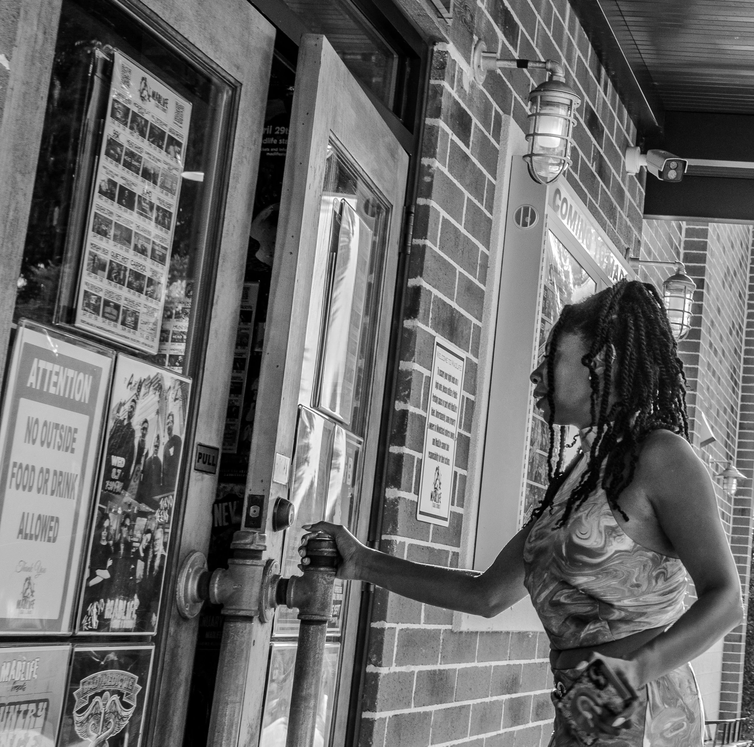 Black and white photo of a woman opening a door to a building. The door has a sign reading 'Attention: No Outside Food or Drink Allowed.' She is wearing a patterned dress and holding a purse.