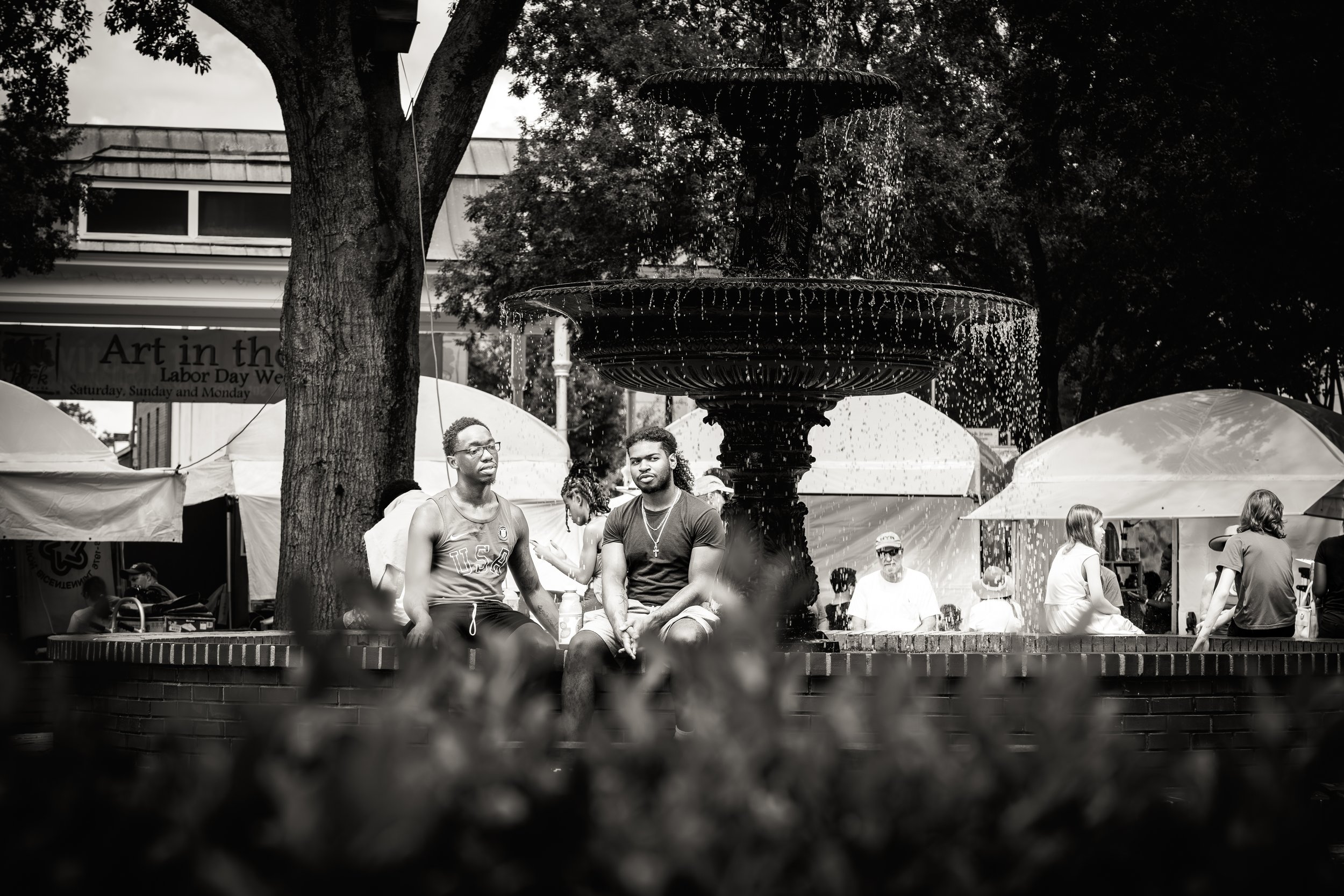 Black and white photo of a public space with a large fountain surrounded by people sitting on its edge, trees, and art event tents in the background.