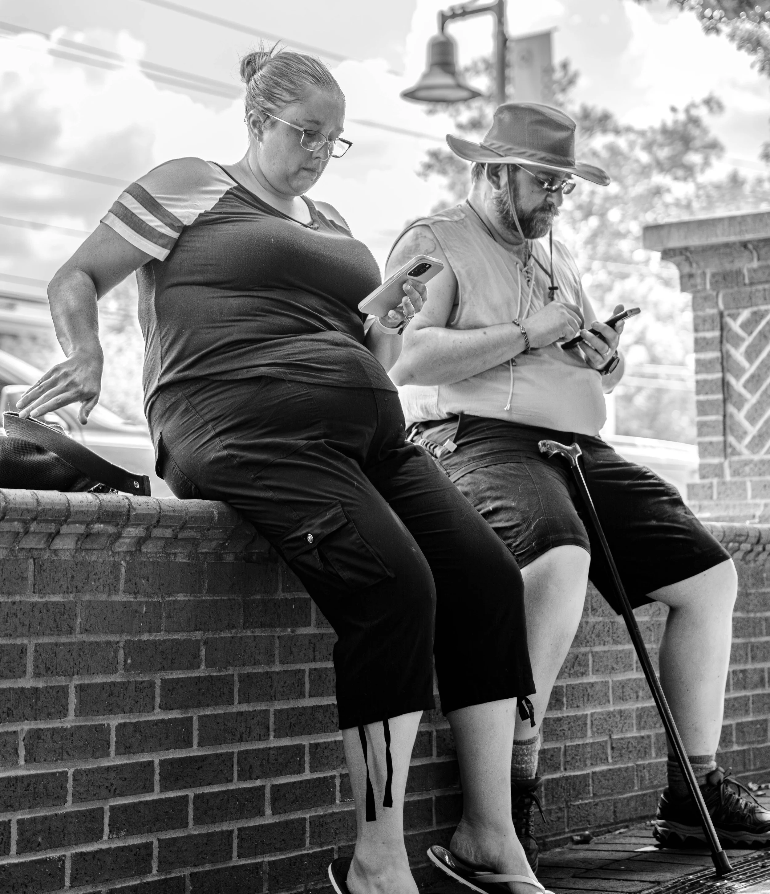 Two people sitting on a brick ledge, each looking at their phones. The person on the left is a woman with glasses and tied-back hair, wearing a short-sleeved shirt. The person on the right is a man with a hat and glasses, holding a cane. Black and wh