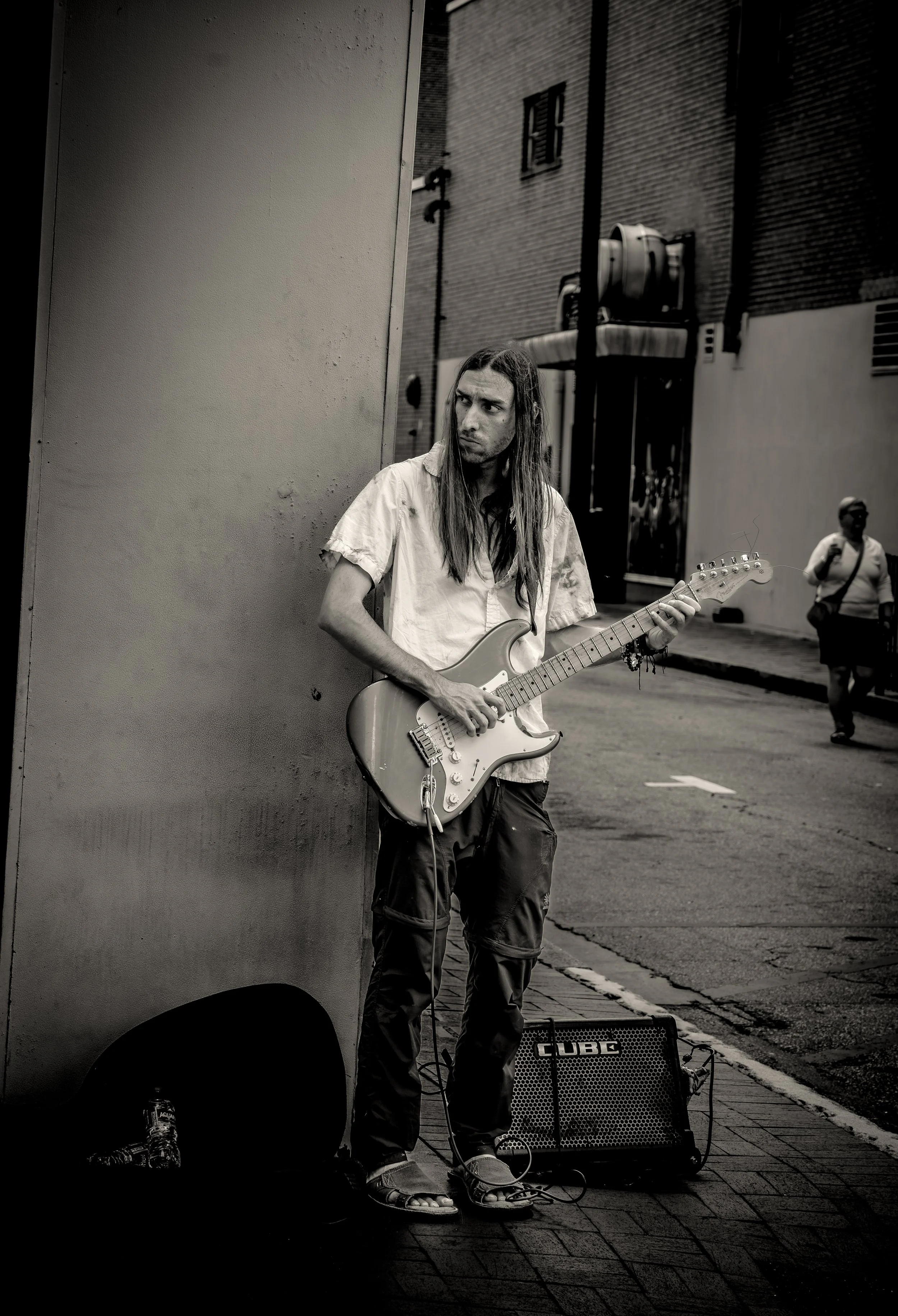 Street musician with long hair playing an electric guitar next to an amplifier on a sidewalk in an urban setting.