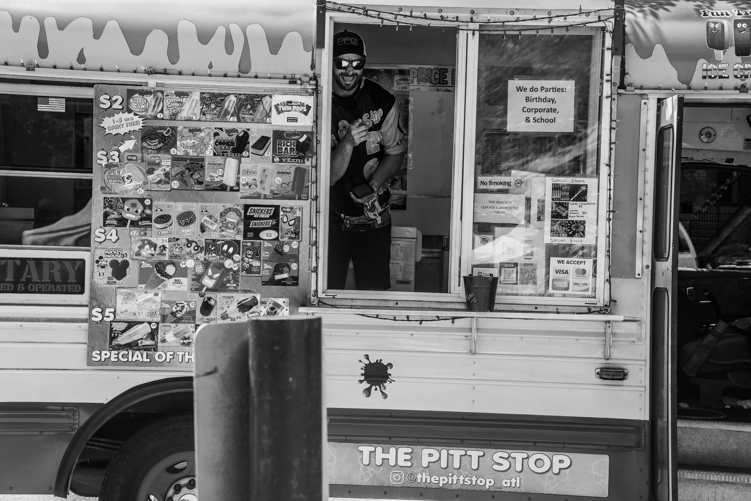 Black and white photo of an ice cream truck with a menu on the side, a person wearing sunglasses smiling from the service window. Sign advertises party services and payment options.