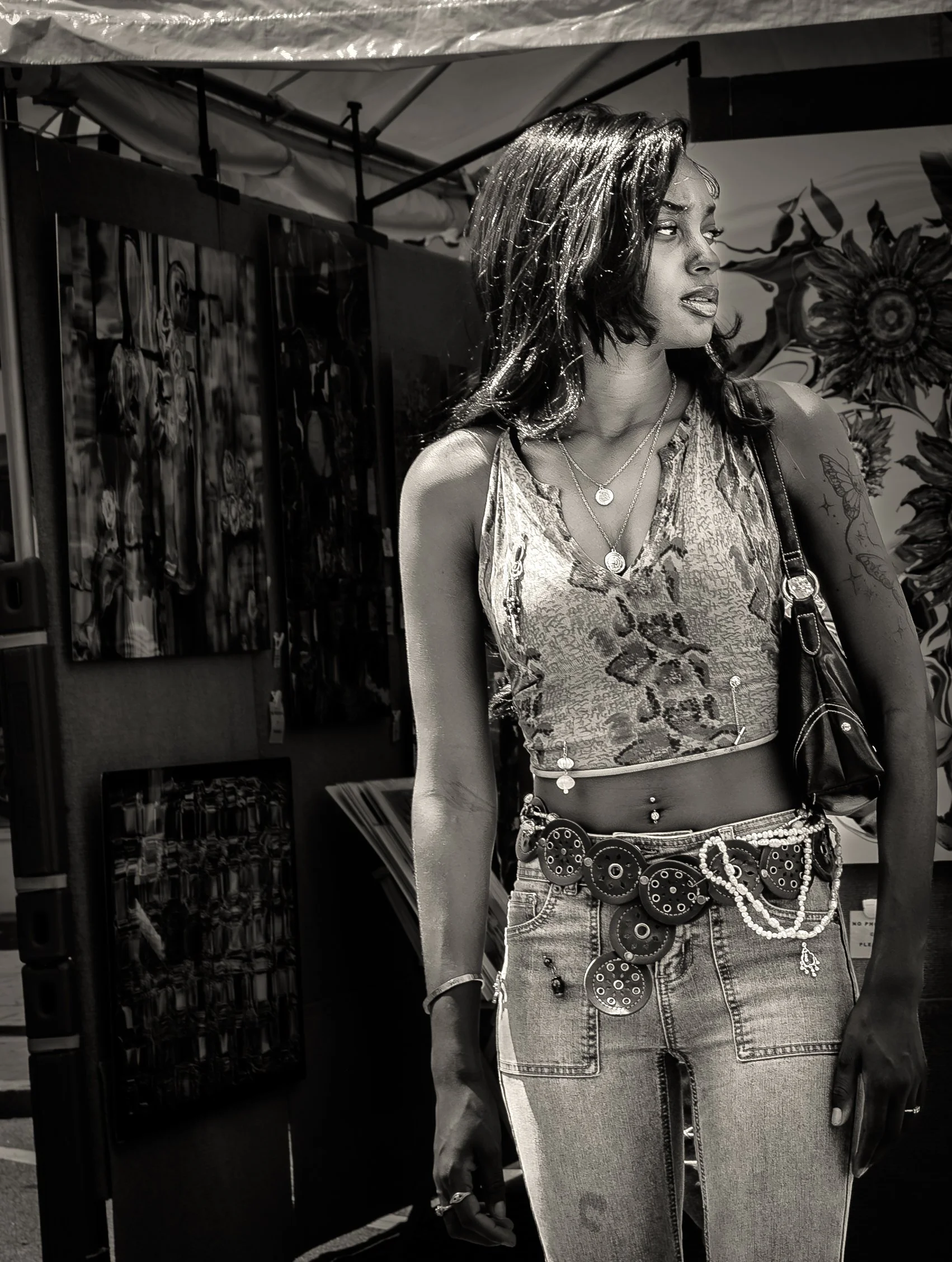 Black and white photo of a person at an art fair, wearing a patterned top and jeans with decorative accessories, standing near art displays under a canopy.
