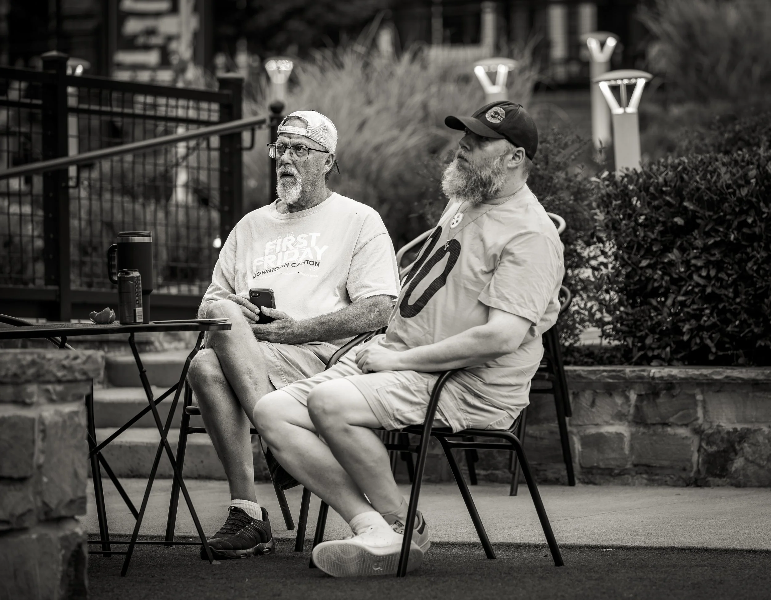 Two men sitting outdoors at a table in a park setting, one wearing a t-shirt and backward cap, and the other in a t-shirt and baseball cap, with a water bottle on the table.