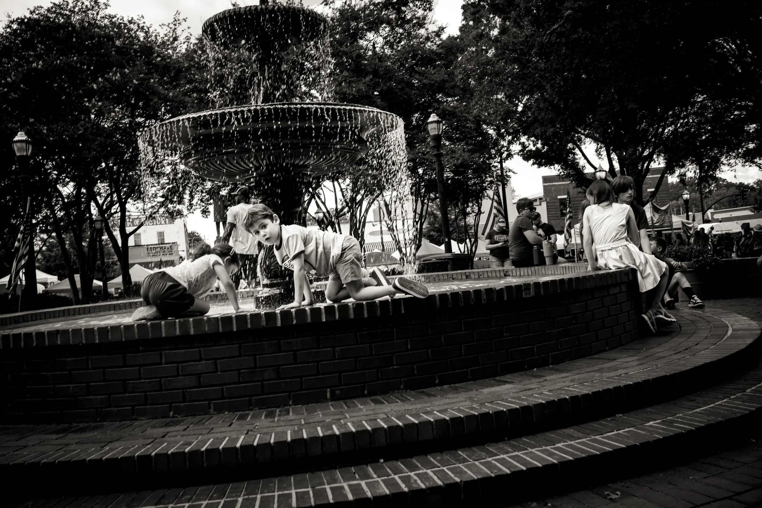 Children playing around a fountain in a park, with trees and people in the background.