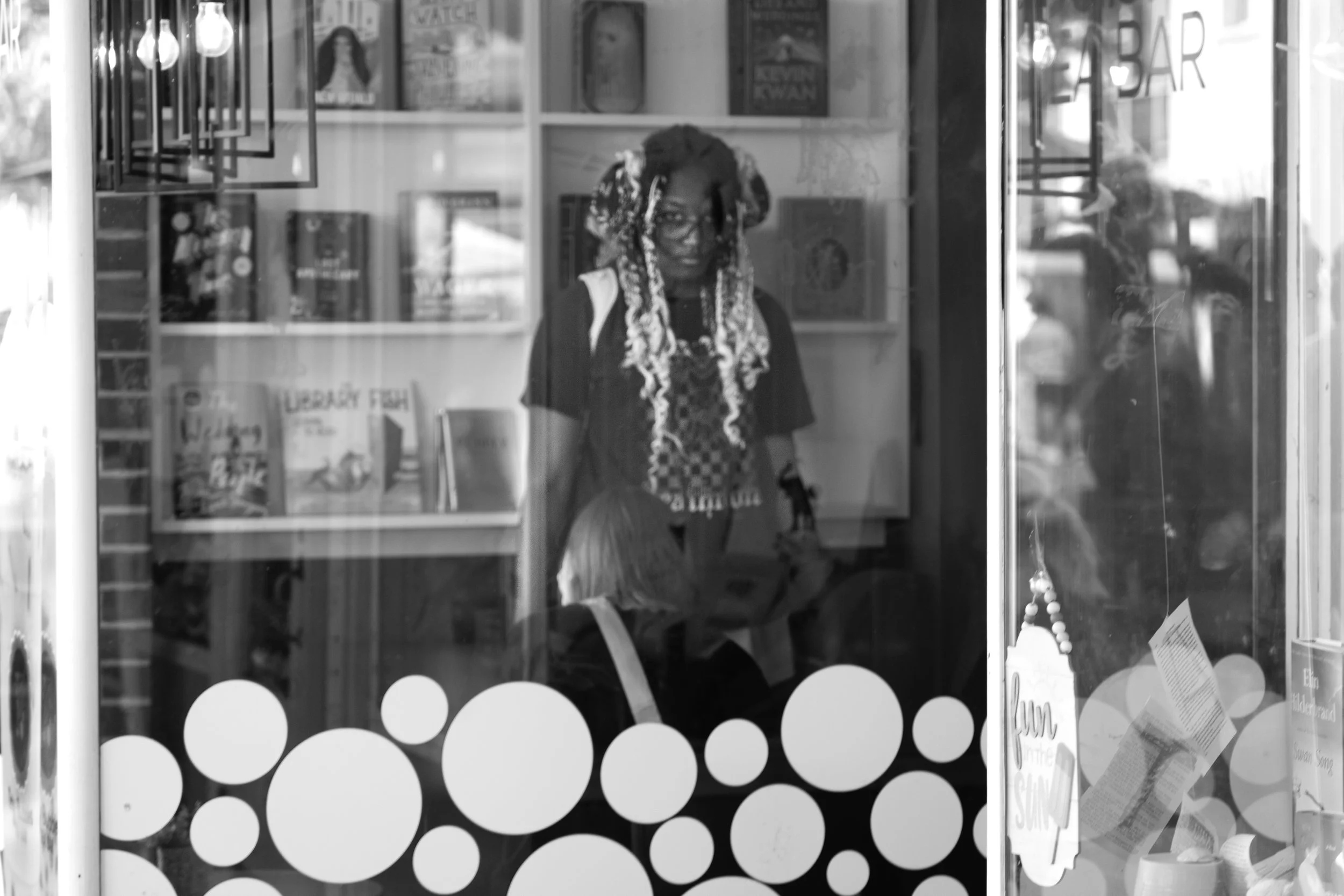Black and white photo of a bookstore window with books on display and circular decals, showing two people inside.